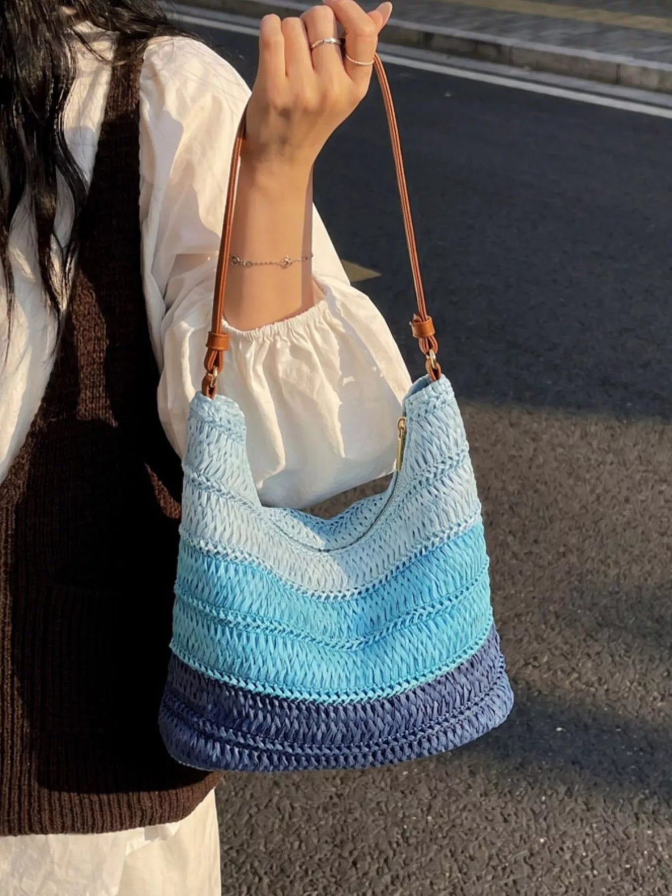 Medium woven straw shoulder bag with blue coastal stripes, curved silhouette, brown shoulder strap, and breezy summer styling.