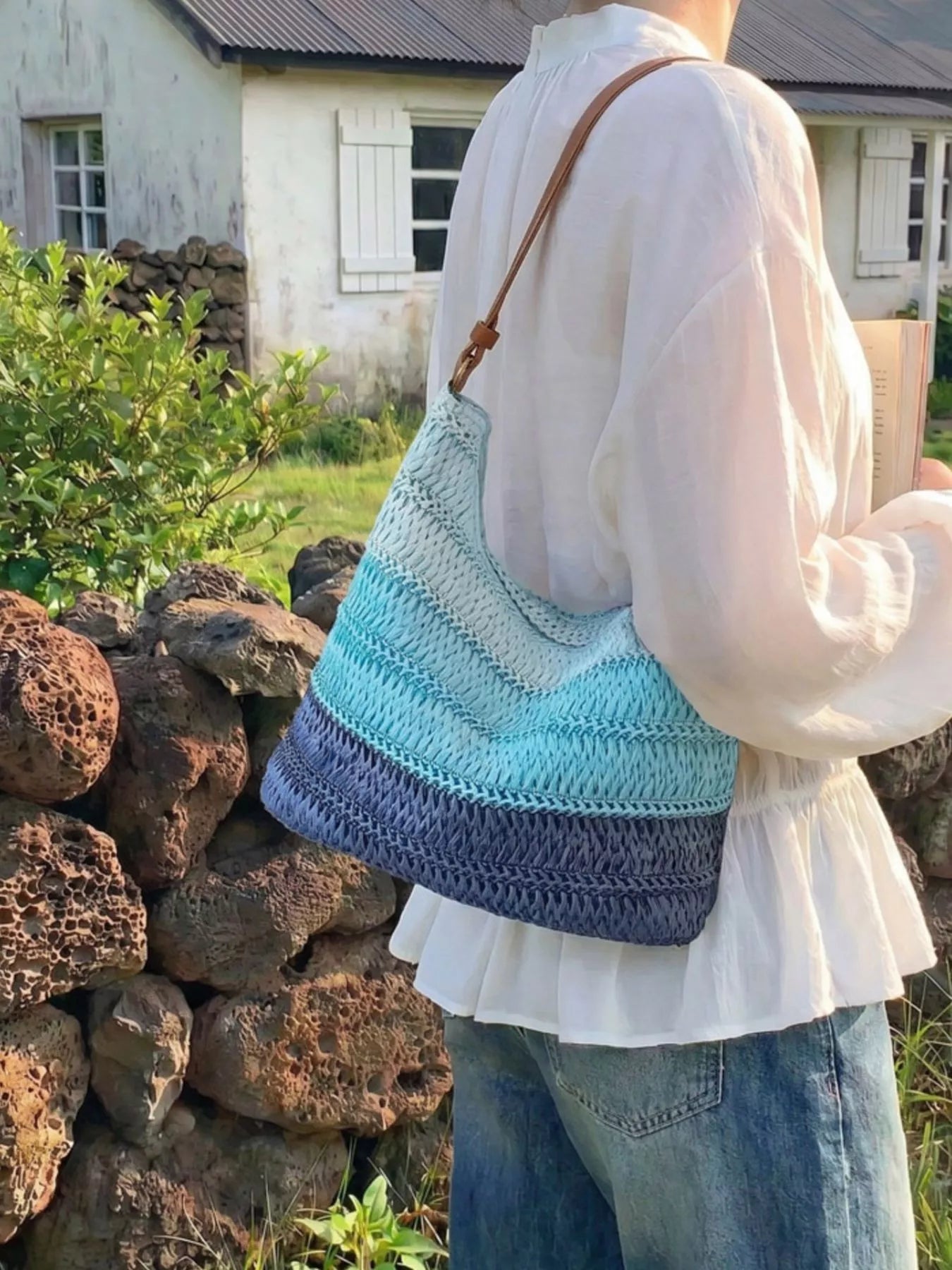 Medium woven straw shoulder bag with blue coastal stripes, curved silhouette, brown shoulder strap, and breezy summer styling.