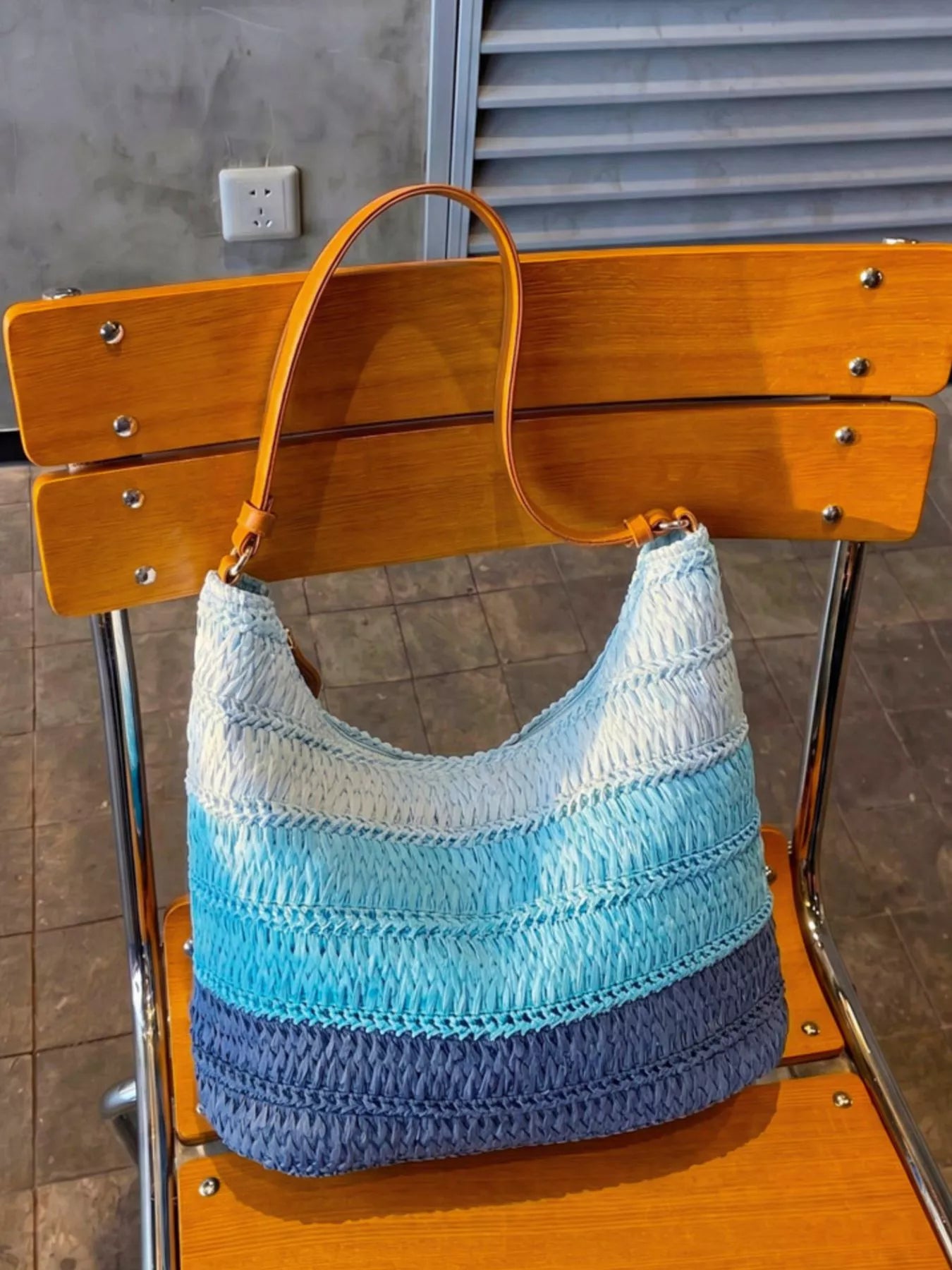 Medium woven straw shoulder bag with blue coastal stripes, curved silhouette, brown shoulder strap, and breezy summer styling.