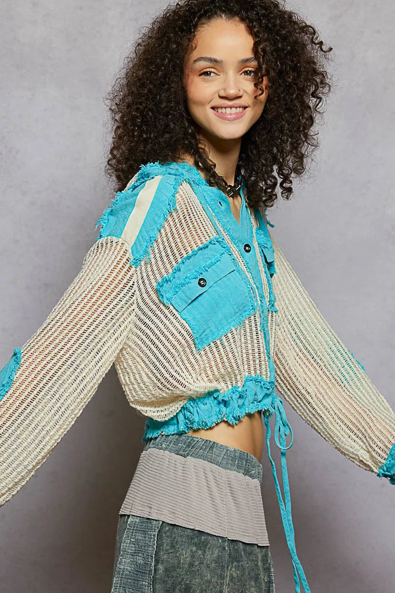 Model wearing an ivory woven hooded button-down top with turquoise contrast patches, cropped drawstring hem, and front pockets.