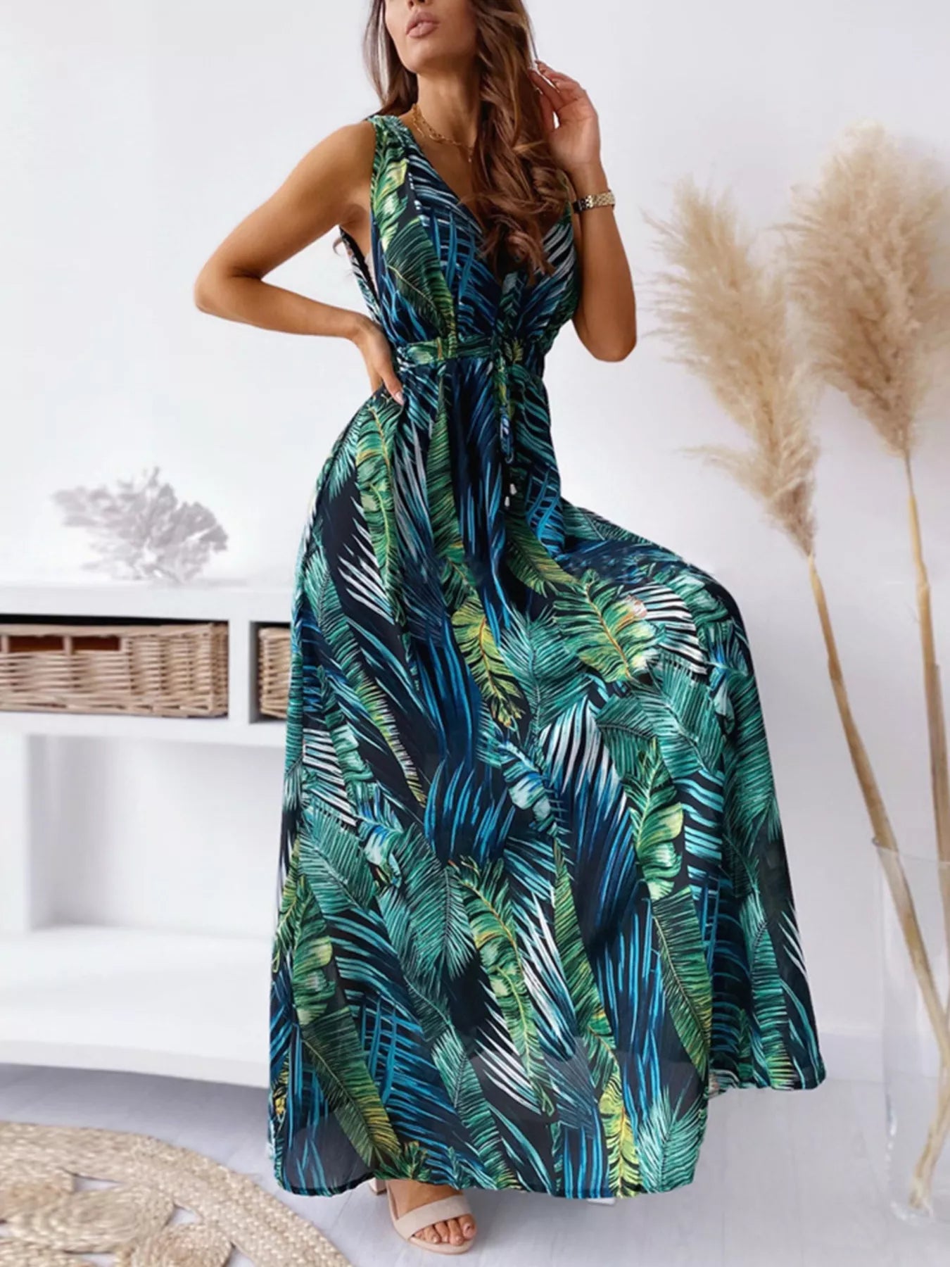 A flowy sleeveless maxi dress with a deep V-neckline and green/blue palm leaf print, styled with a drawstring waist.