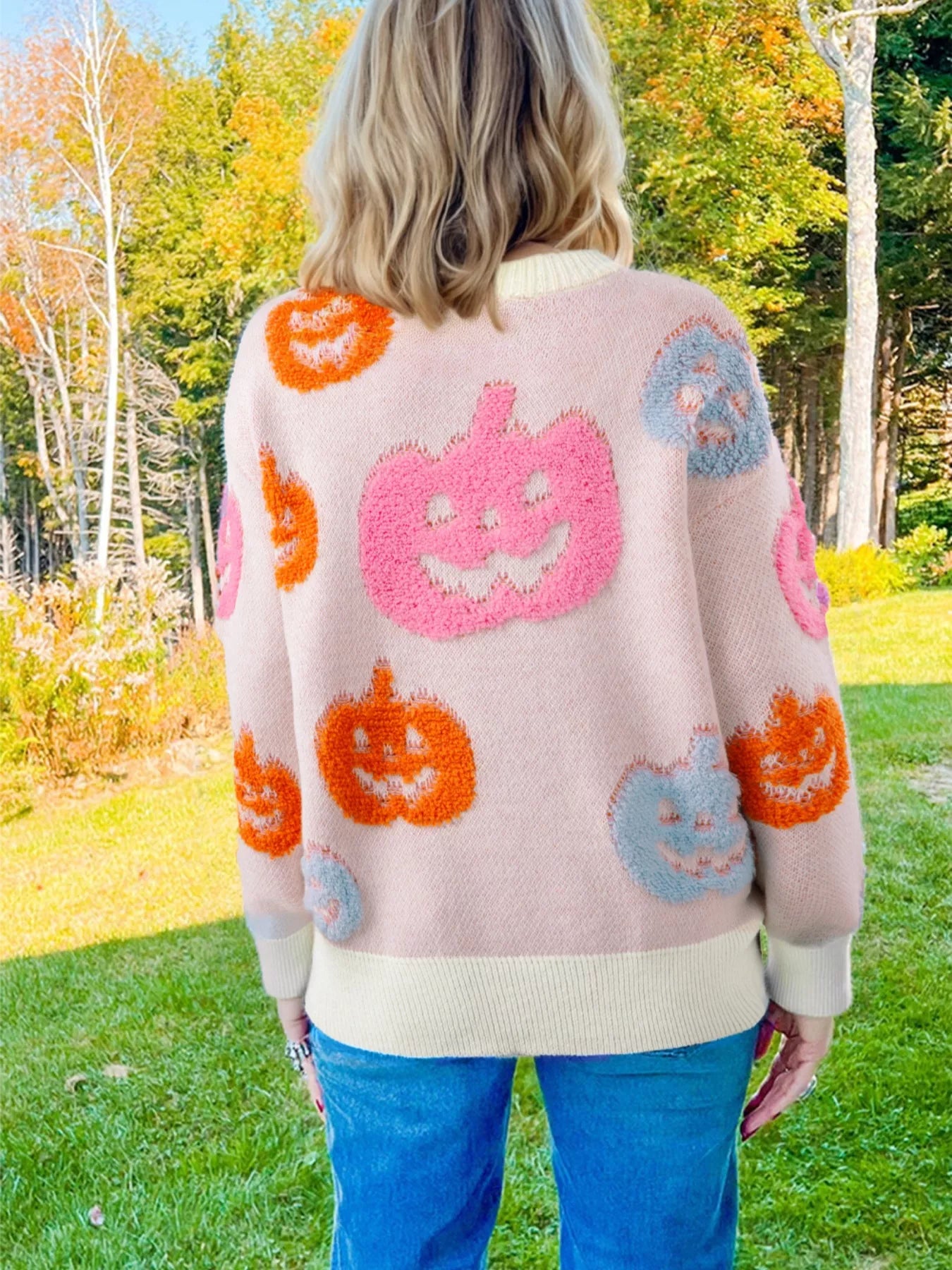 Woman wearing a cream sweater decorated with pastel orange, pink, and blue textured jack-o’-lanterns — cozy Halloween sweater from A Whimsical Affair.