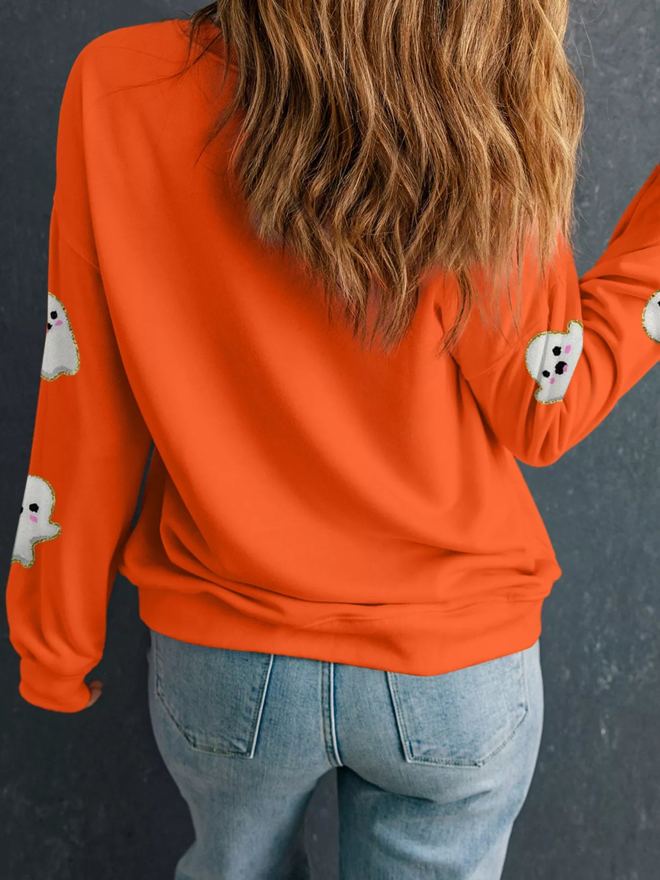 Woman wearing a bright orange sweatshirt decorated with cute white ghosts outlined in gold — festive Halloween sweatshirt from A Whimsical Affair.