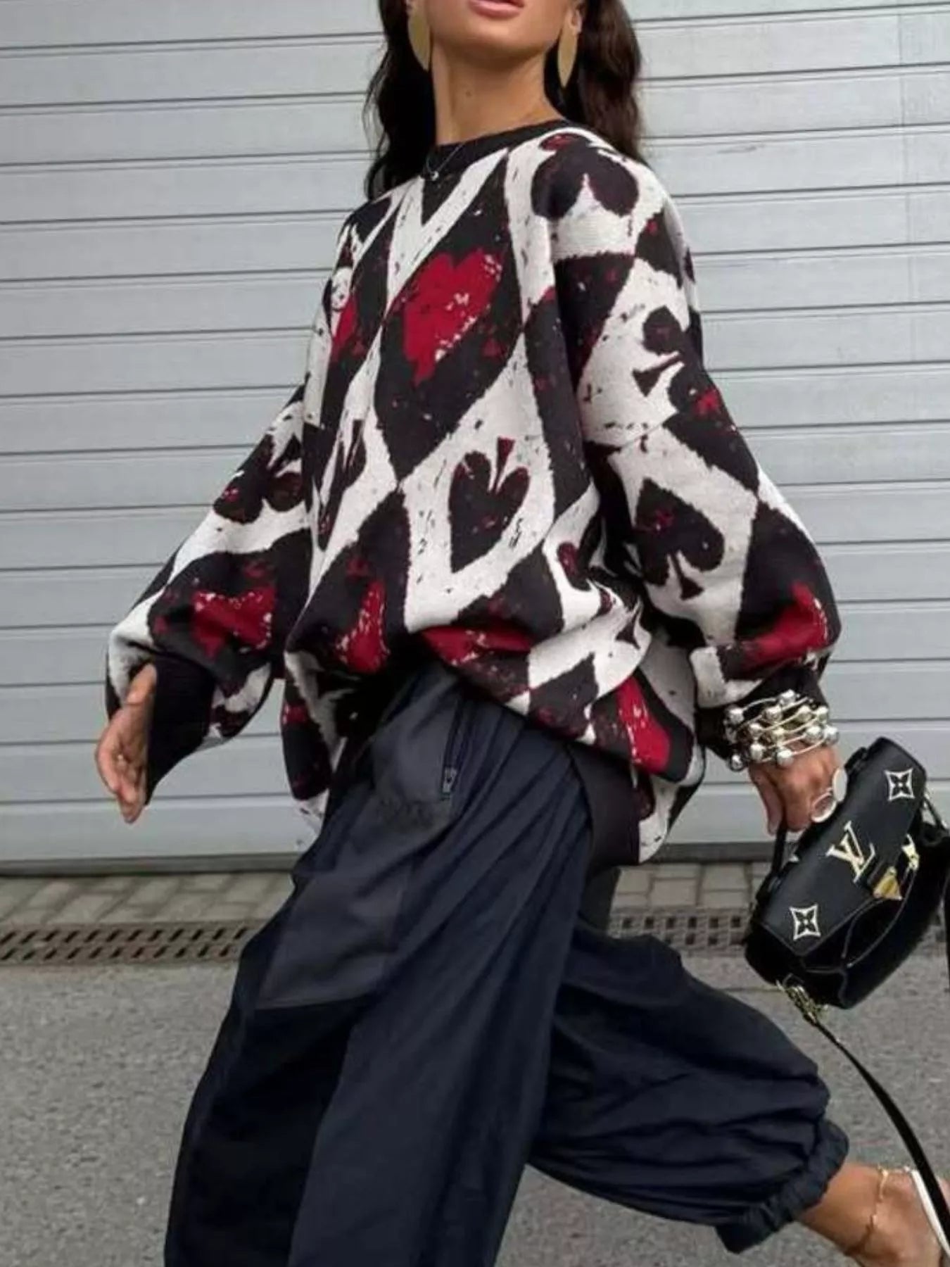 Oversized sweater with red and black playing card motif in bold diamond and spade pattern, styled with black pants and heels.