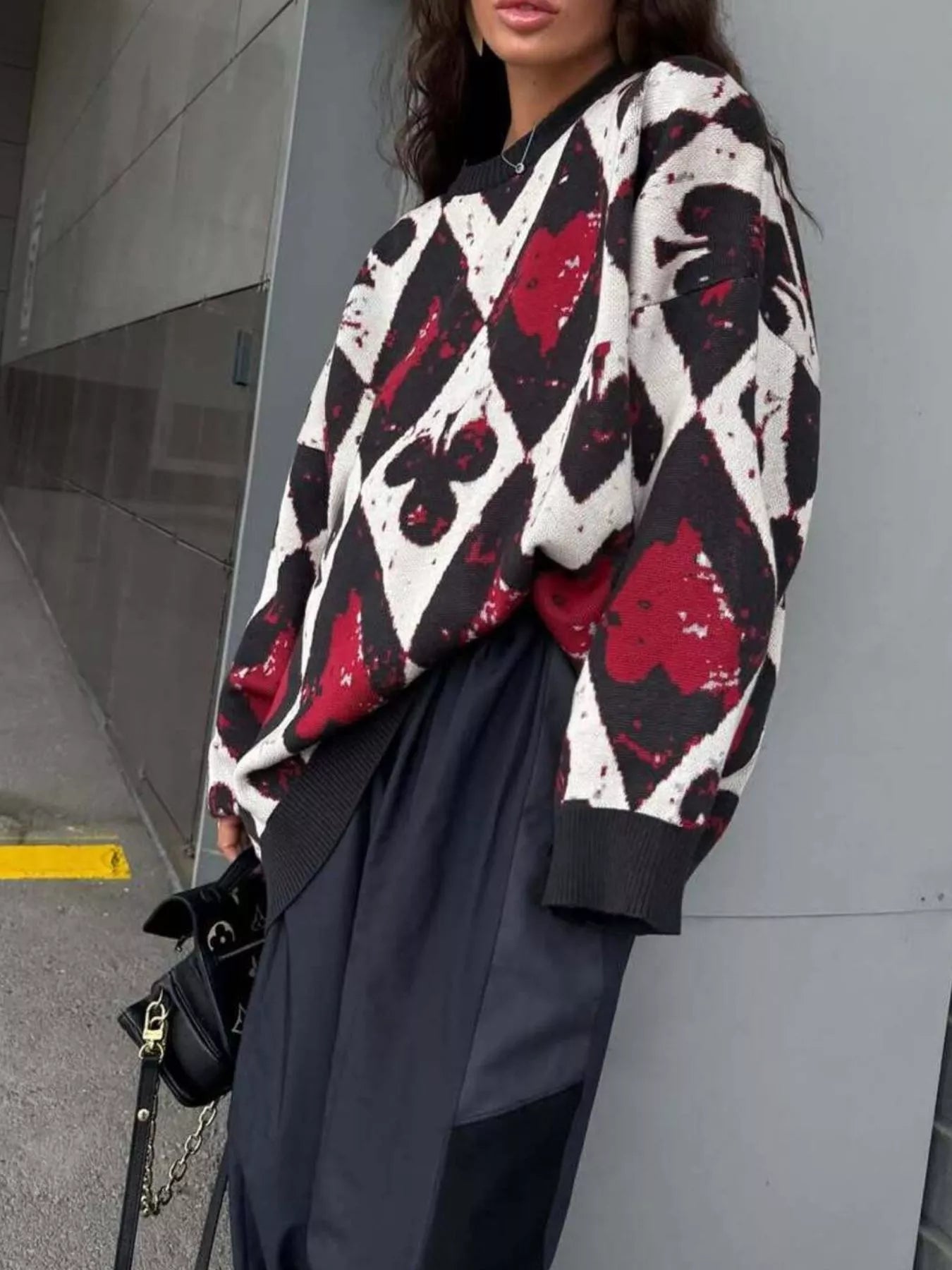 Oversized sweater with red and black playing card motif in bold diamond and spade pattern, styled with black pants and heels.
