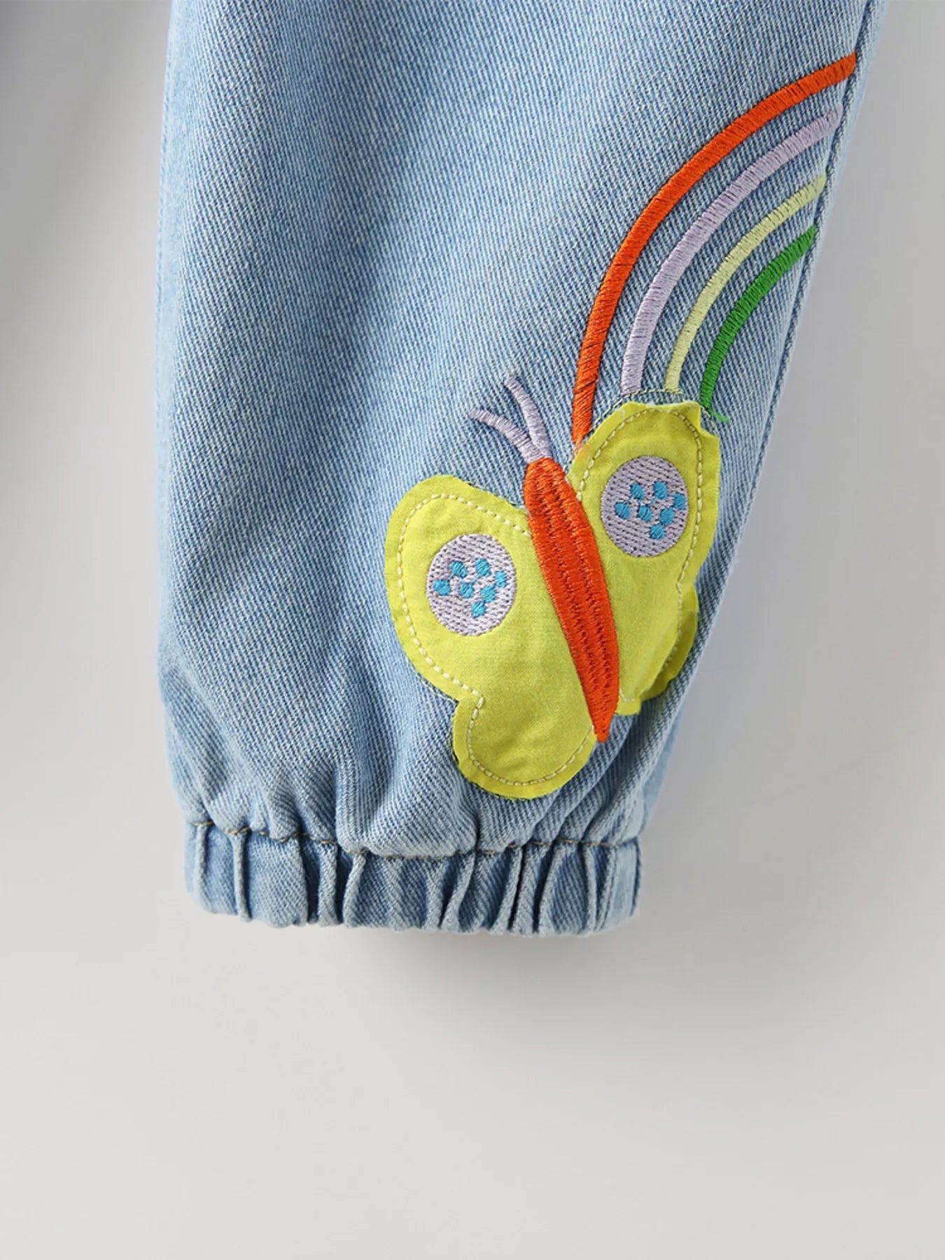 Light-wash kids' denim joggers with embroidered rainbow, bee, and butterfly details. Elastic waist and pockets for comfy, playful style.