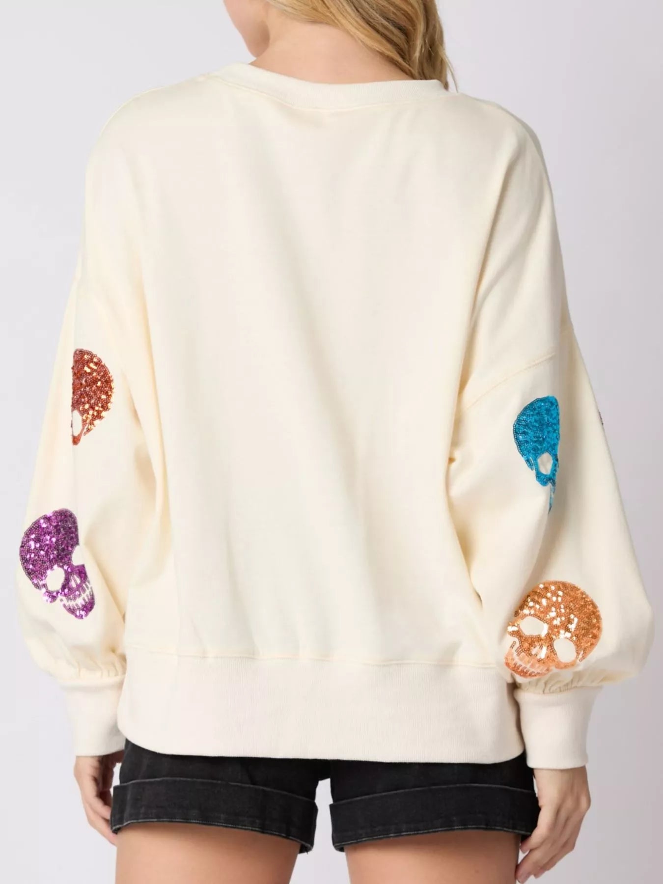 Woman wearing a cream sweatshirt with rainbow-colored sequin skulls — cozy and bold Halloween pullover from A Whimsical Affair.