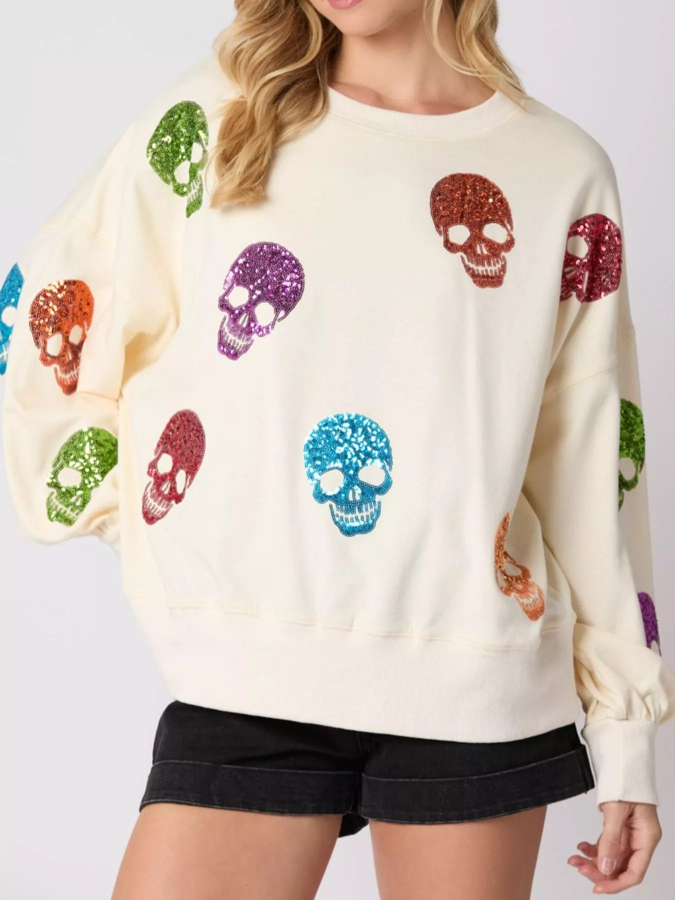 Woman wearing a cream sweatshirt with rainbow-colored sequin skulls — cozy and bold Halloween pullover from A Whimsical Affair.