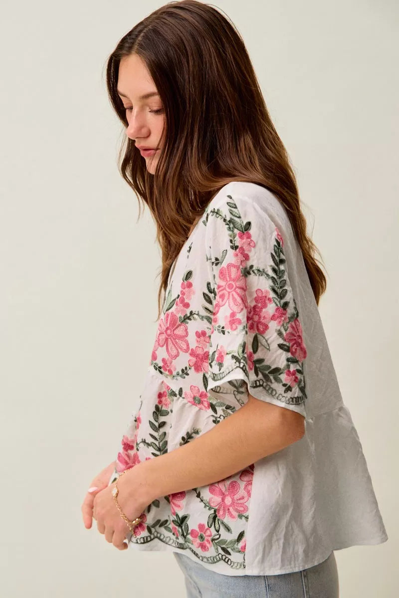 White boho blouse with pink floral embroidery, green vine details, V-neckline, short sleeves, and relaxed flowy fit styled with jeans.