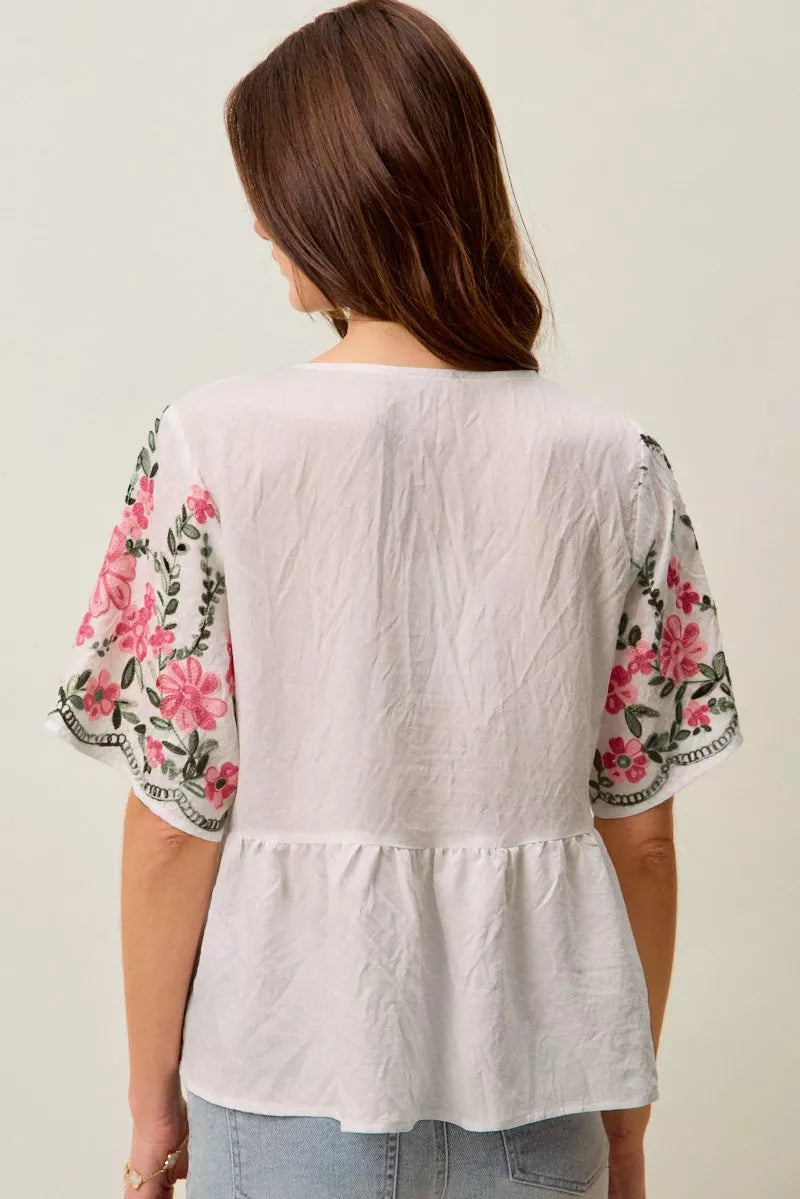 White boho blouse with pink floral embroidery, green vine details, V-neckline, short sleeves, and relaxed flowy fit styled with jeans.