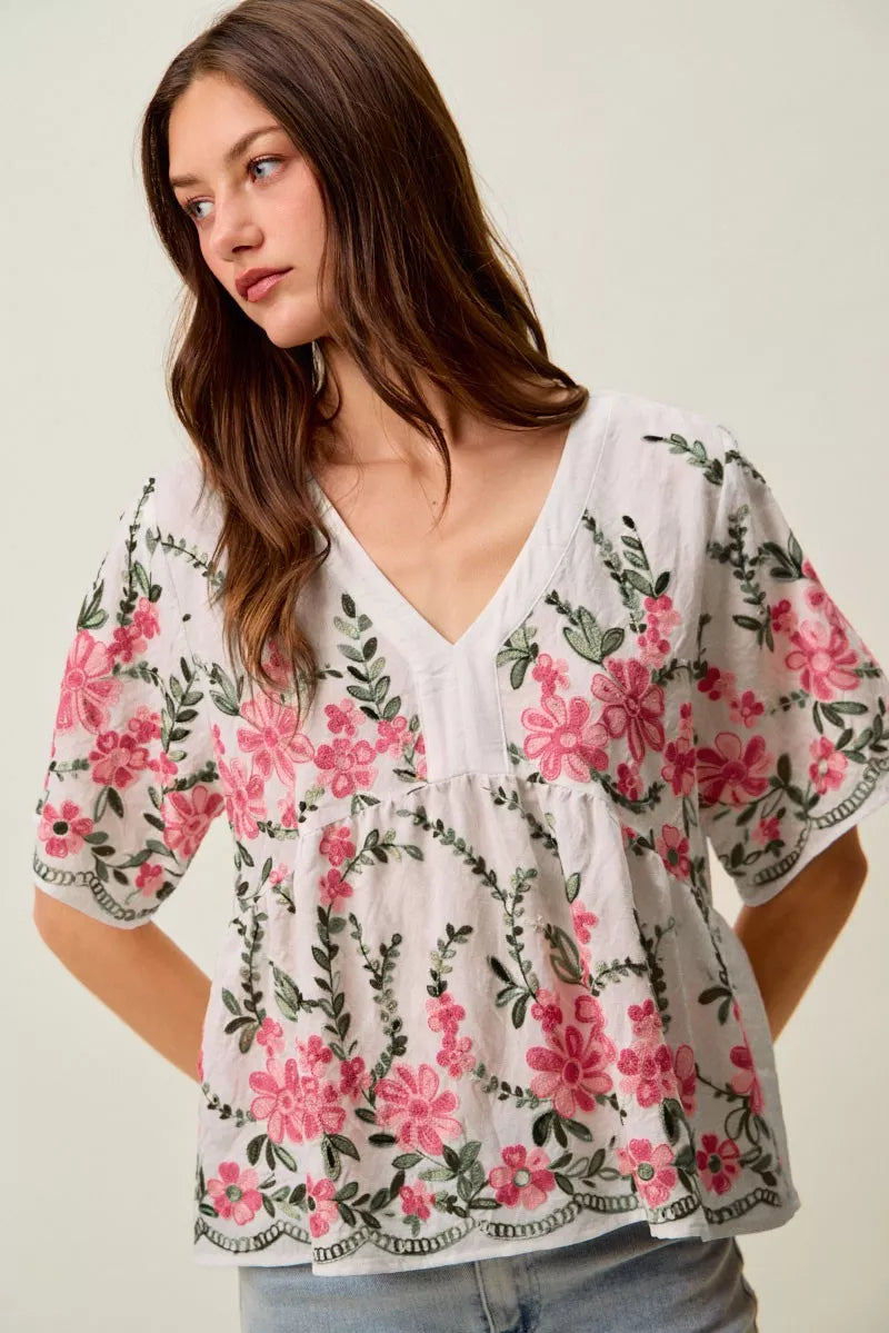 White boho blouse with pink floral embroidery, green vine details, V-neckline, short sleeves, and relaxed flowy fit styled with jeans.