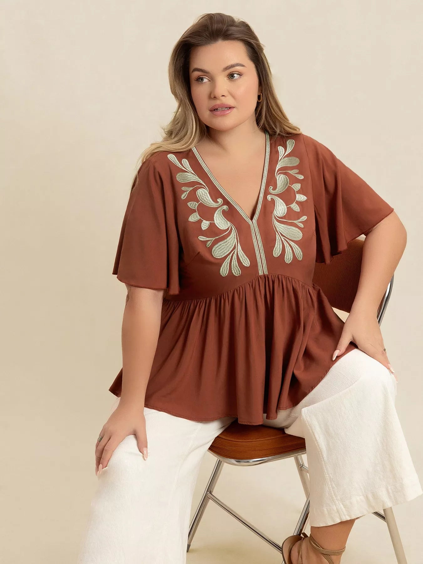 A woman wearing a rust-colored V-neck plus size blouse with embroidered floral detailing near the neckline, short flutter sleeves, and a ruched waist design.