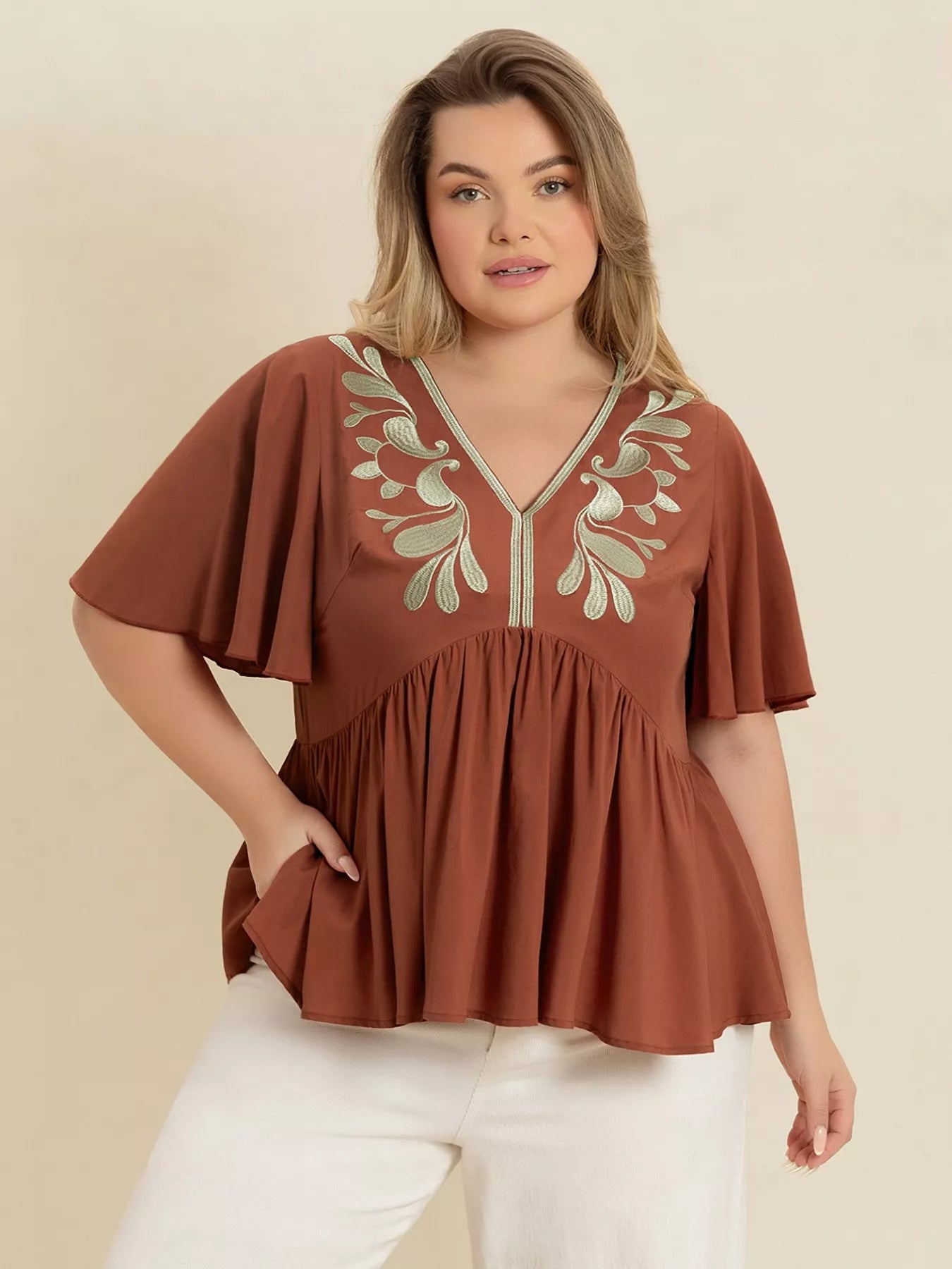 A woman wearing a rust-colored V-neck plus size blouse with embroidered floral detailing near the neckline, short flutter sleeves, and a ruched waist design.