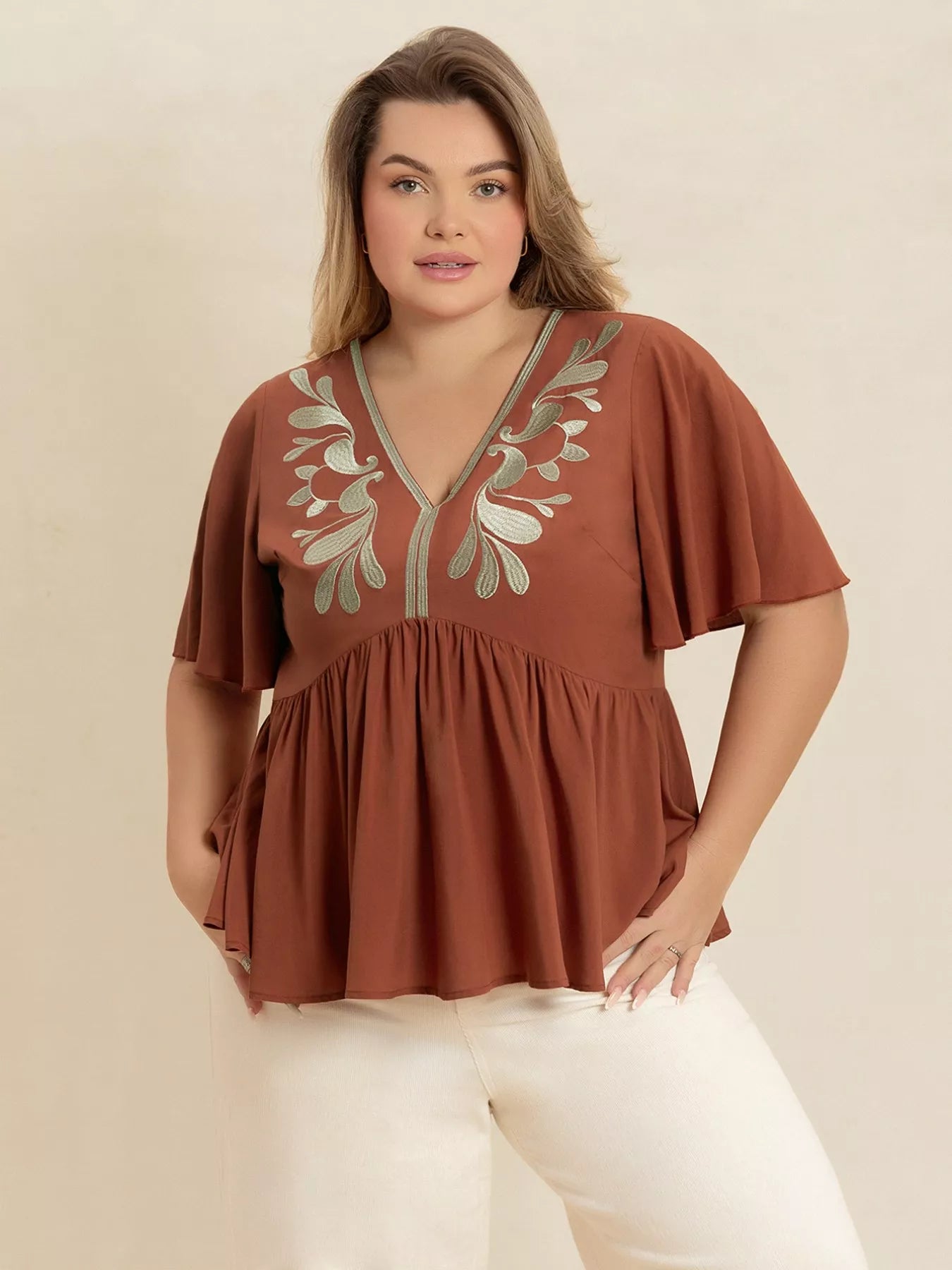 A woman wearing a rust-colored V-neck plus size blouse with embroidered floral detailing near the neckline, short flutter sleeves, and a ruched waist design.