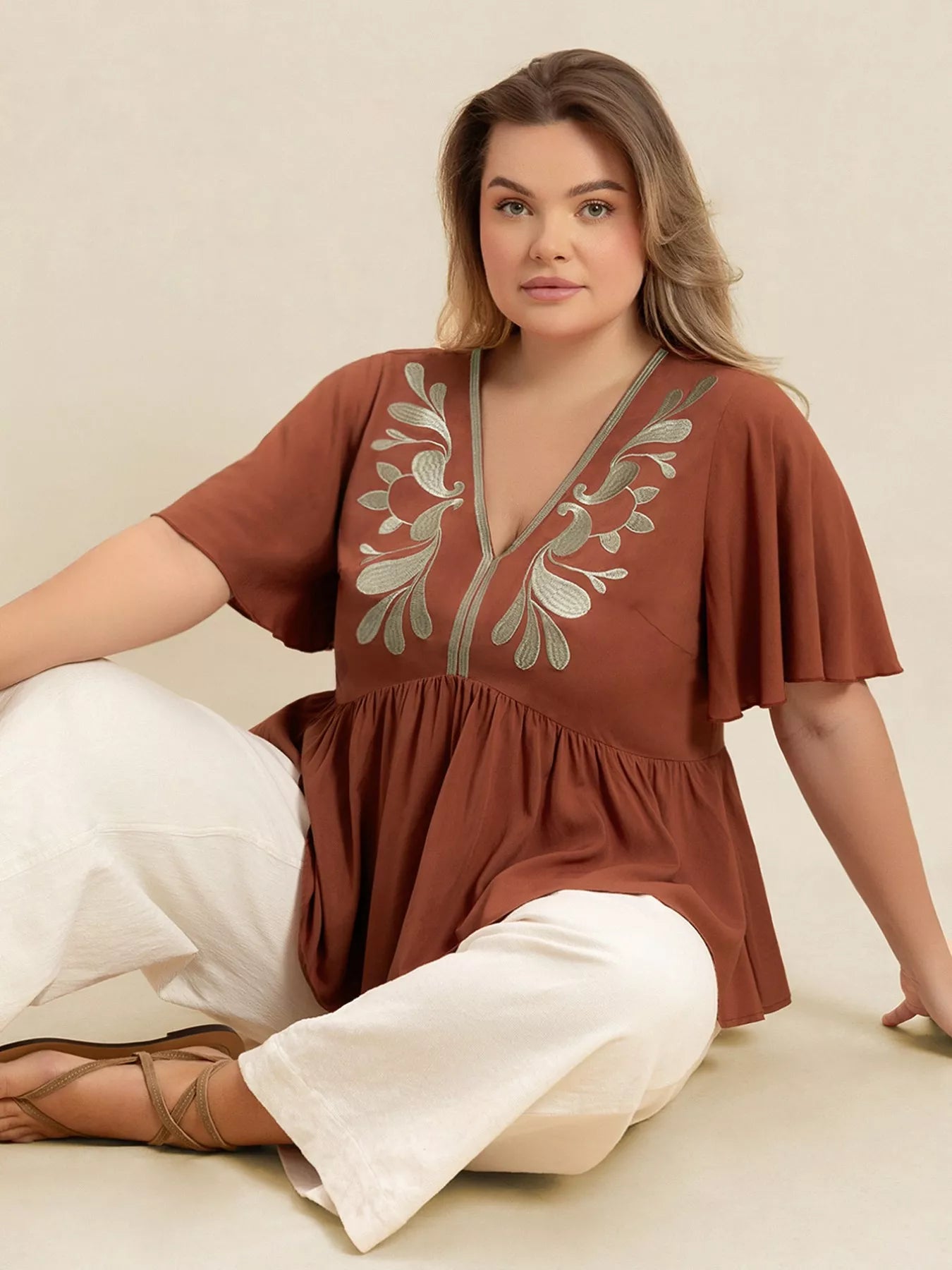 A woman wearing a rust-colored V-neck plus size blouse with embroidered floral detailing near the neckline, short flutter sleeves, and a ruched waist design.