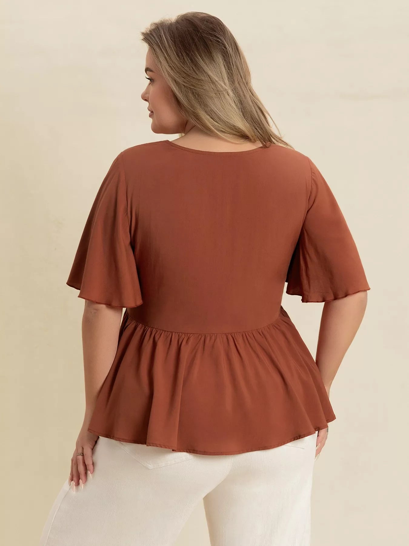 A woman wearing a rust-colored V-neck plus size blouse with embroidered floral detailing near the neckline, short flutter sleeves, and a ruched waist design.