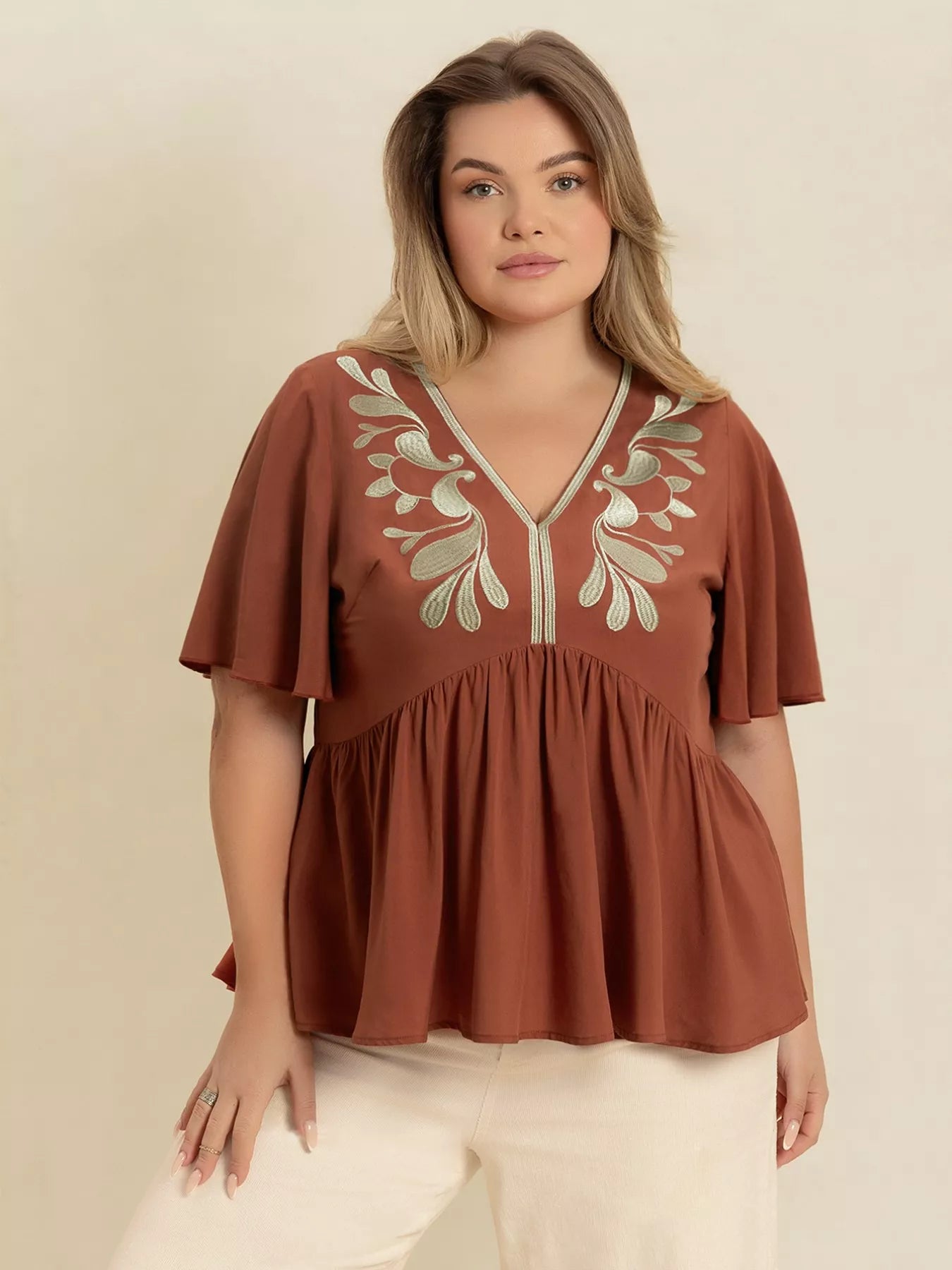 A woman wearing a rust-colored V-neck plus size blouse with embroidered floral detailing near the neckline, short flutter sleeves, and a ruched waist design.