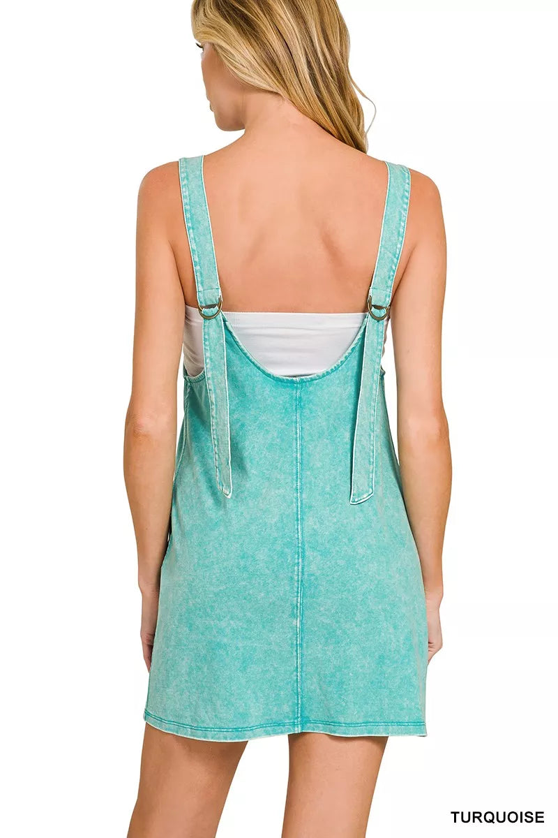 Model wearing a seafoam acid-washed overall mini dress with a sleeveless silhouette and front pockets.
