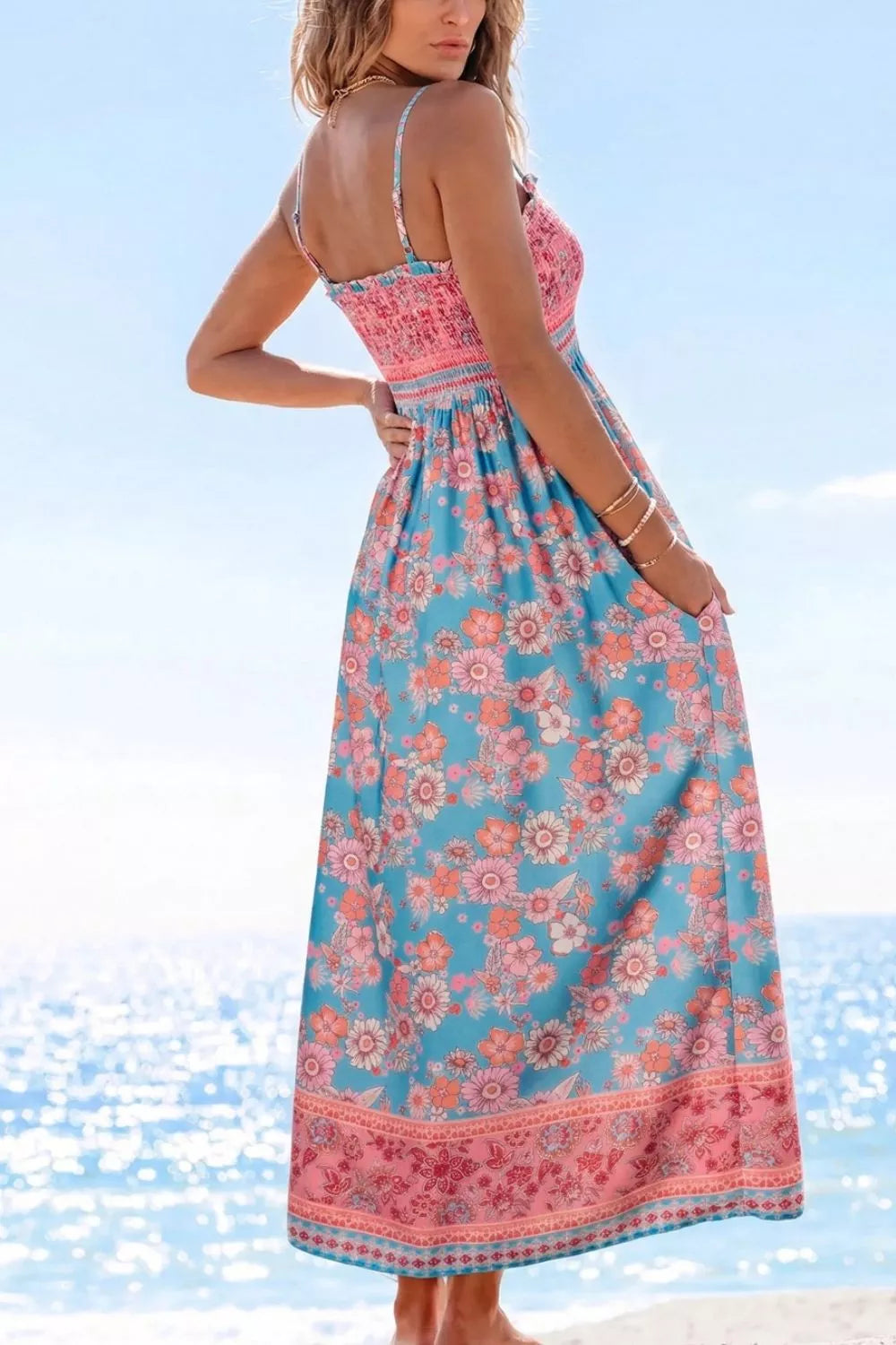 Blue floral smocked maxi dress with spaghetti straps, pink border detailing, and hidden pockets worn on the beach.