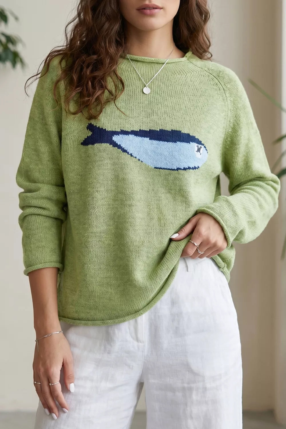 Green knit sweater with a blue fish graphic, ribbed detail, long sleeves, and relaxed slightly stretchy fit styled with white pants.