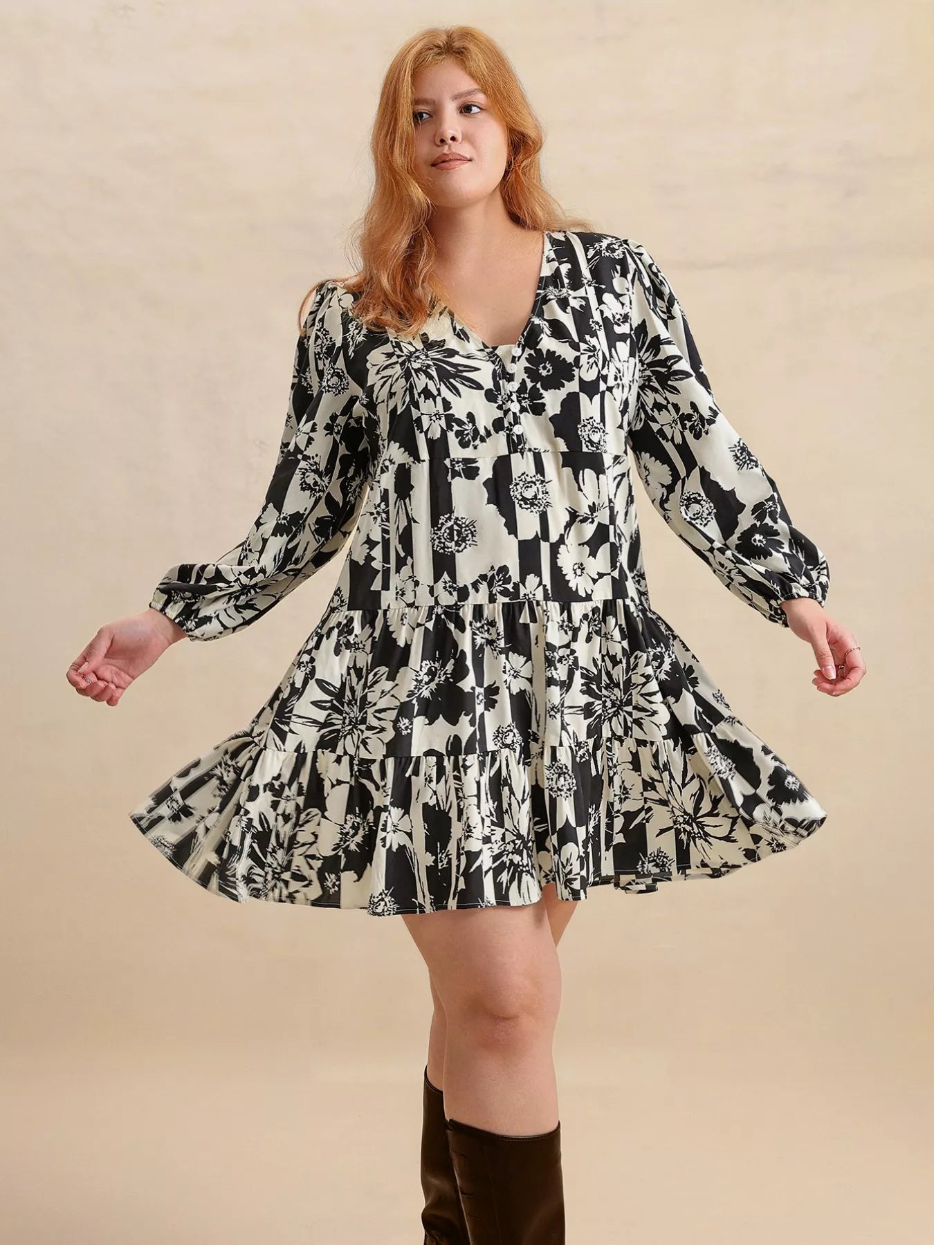 Plus size model wearing the Shadow Petals Mini Dress, a black-and-white floral print mini dress with long sleeves, ruched details, and a flowy hemline, styled with knee-high boots.