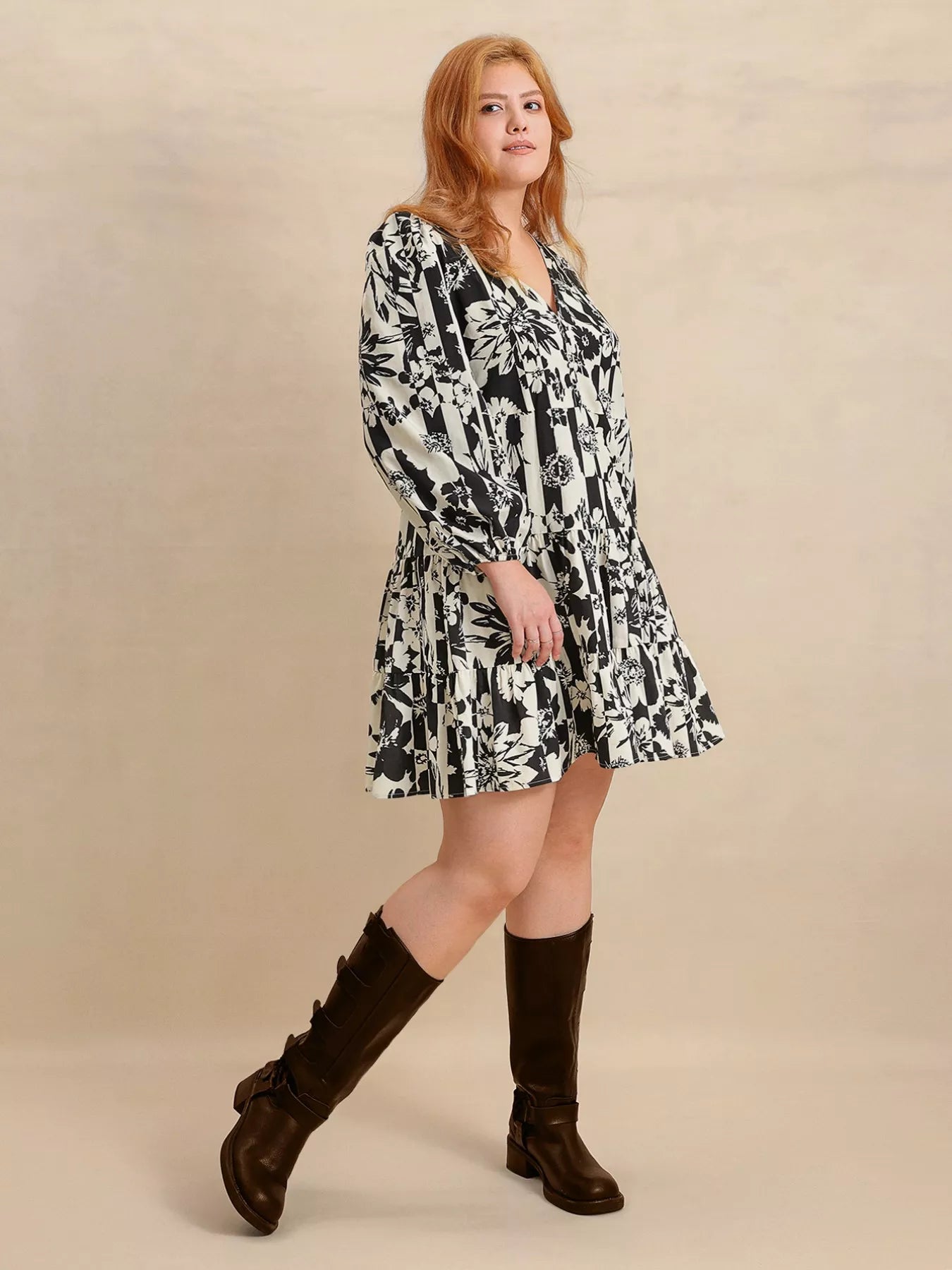 Plus size model wearing the Shadow Petals Mini Dress, a black-and-white floral print mini dress with long sleeves, ruched details, and a flowy hemline, styled with knee-high boots.