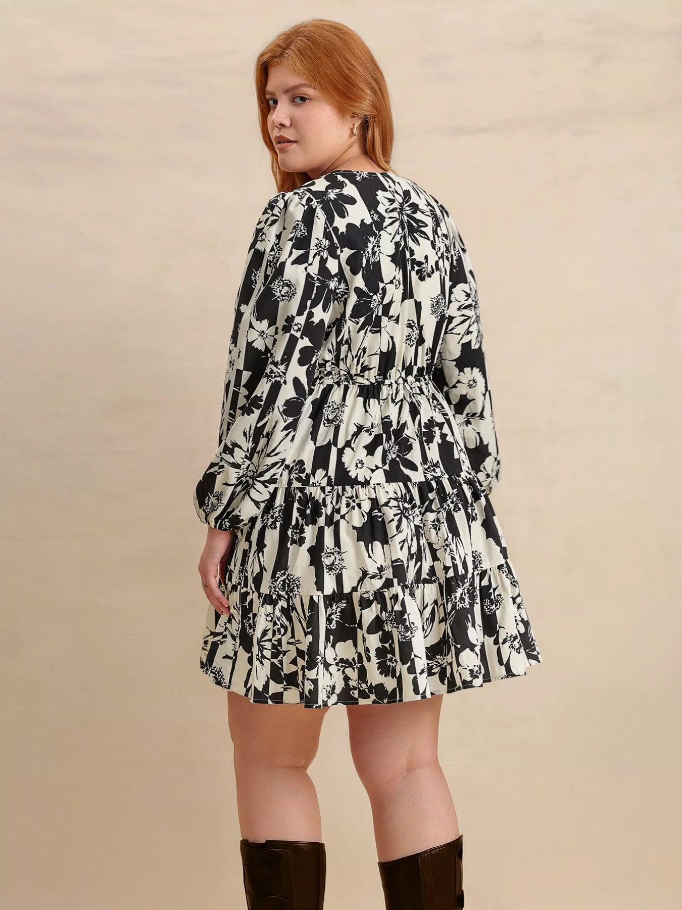 Plus size model wearing the Shadow Petals Mini Dress, a black-and-white floral print mini dress with long sleeves, ruched details, and a flowy hemline, styled with knee-high boots.