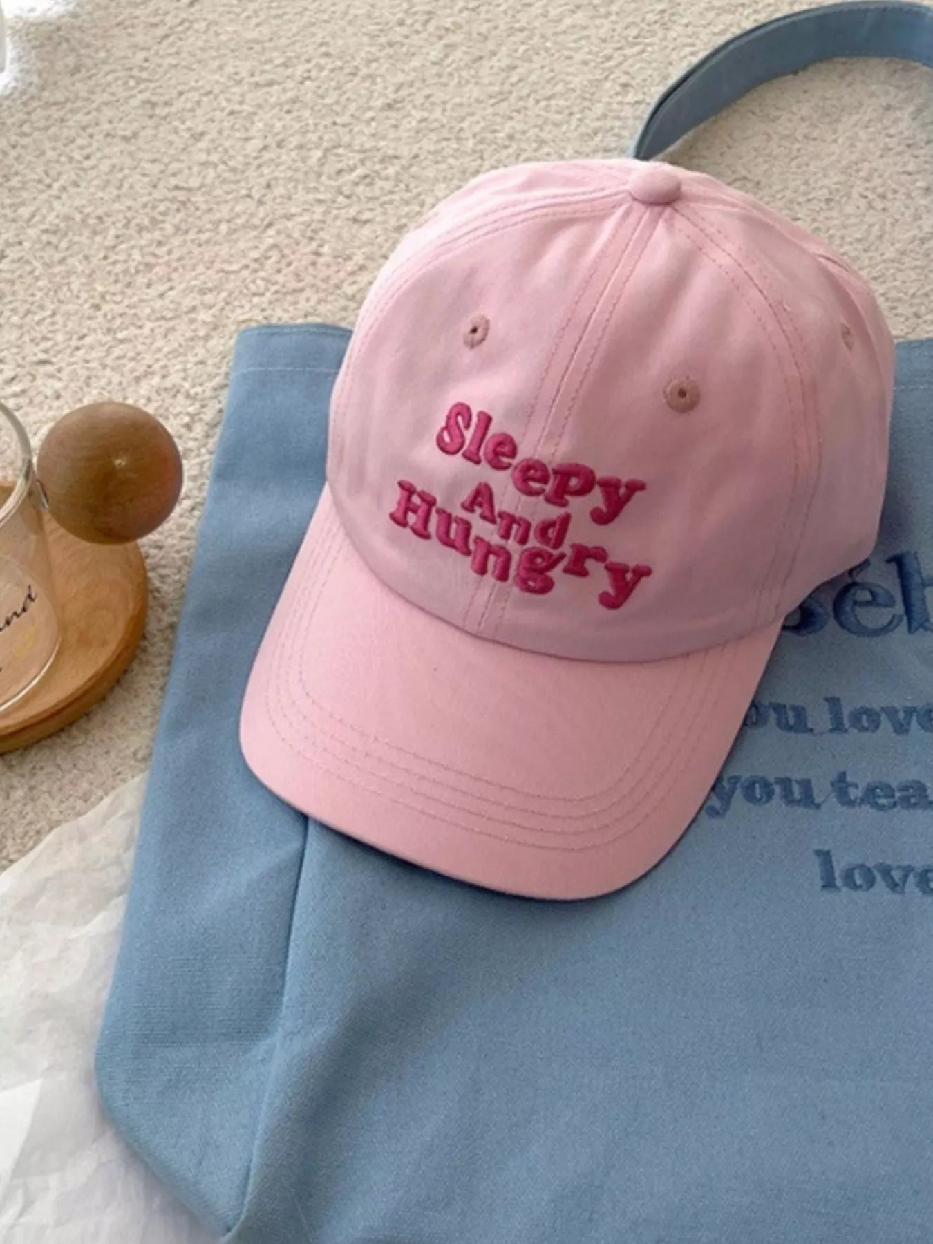 cotton baseball cap with colorful embroidered “sleepy and hungry” lettering on the front, shown on a light blue tote bag.