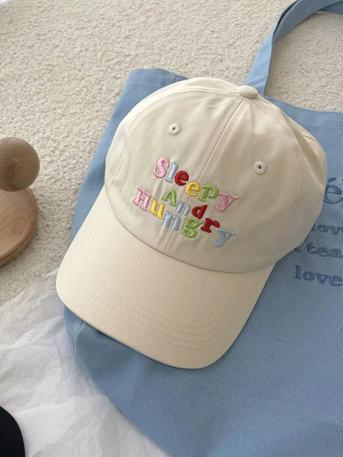 cotton baseball cap with colorful embroidered “sleepy and hungry” lettering on the front, shown on a light blue tote bag.