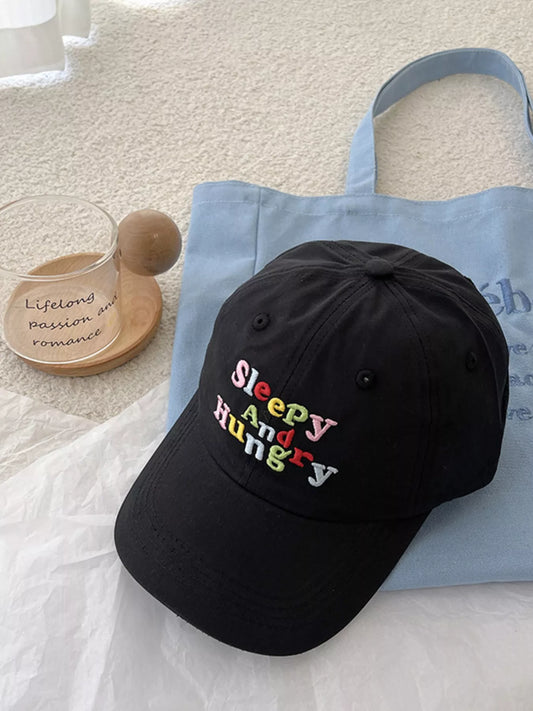 cotton baseball cap with colorful embroidered “sleepy and hungry” lettering on the front, shown on a light blue tote bag.