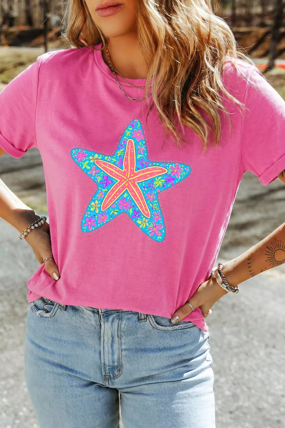 Bright pink short sleeve graphic tee with a colorful floral starfish design on the front, styled with light wash denim jeans.