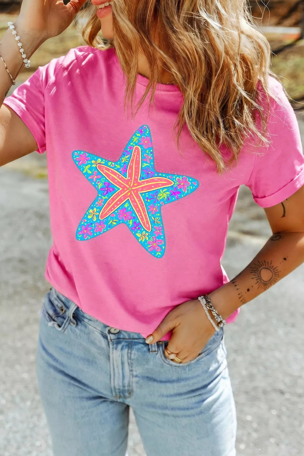 Bright pink short sleeve graphic tee with a colorful floral starfish design on the front, styled with light wash denim jeans.