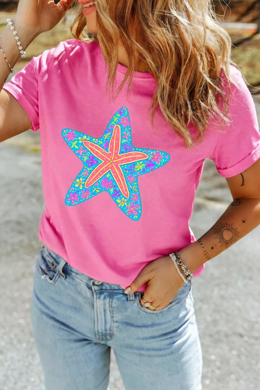 Bright pink short sleeve graphic tee with a colorful floral starfish design on the front, styled with light wash denim jeans.
