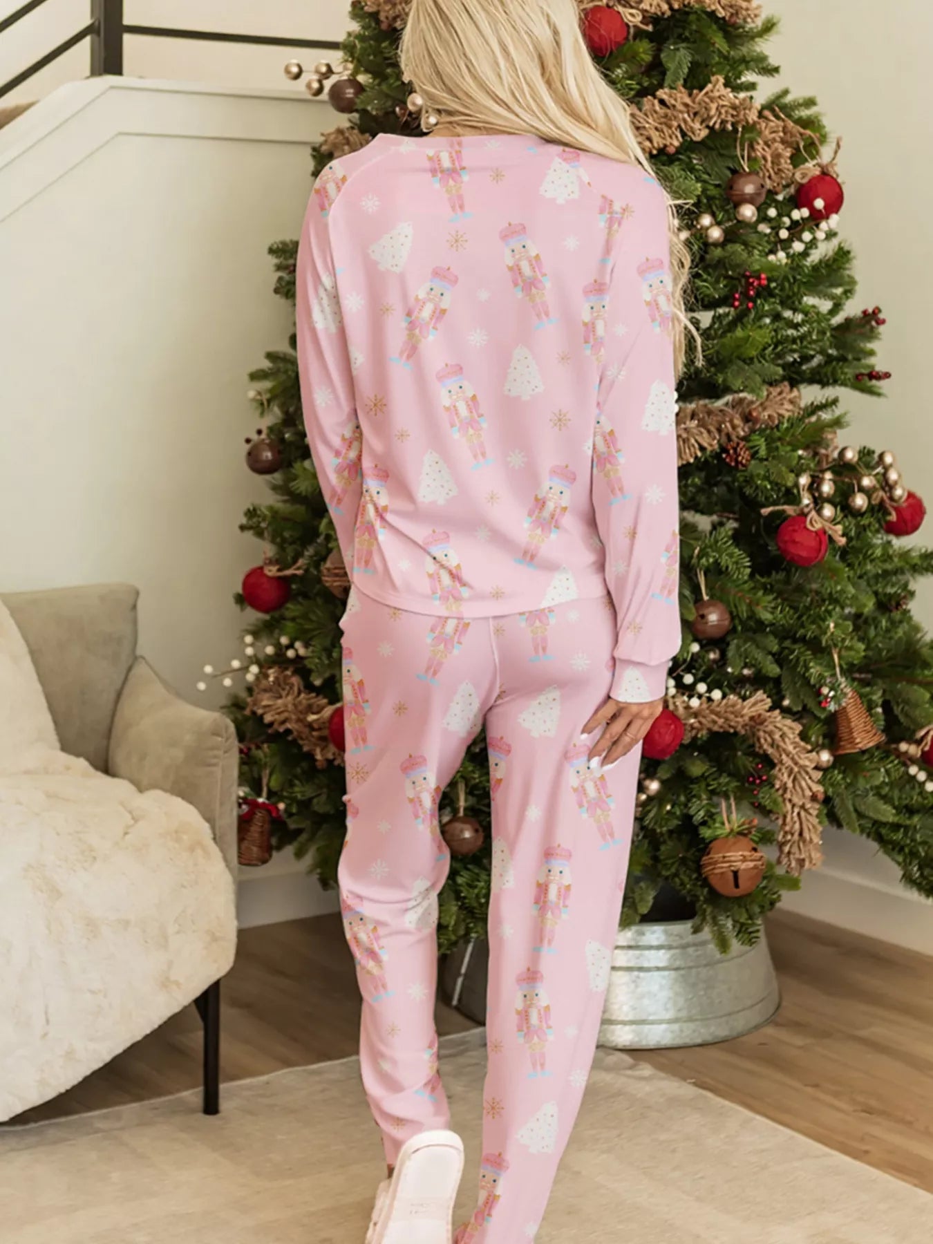Pink two-piece nutcracker-print lounge set with long sleeves and drawstring pants worn indoors beside a Christmas tree.