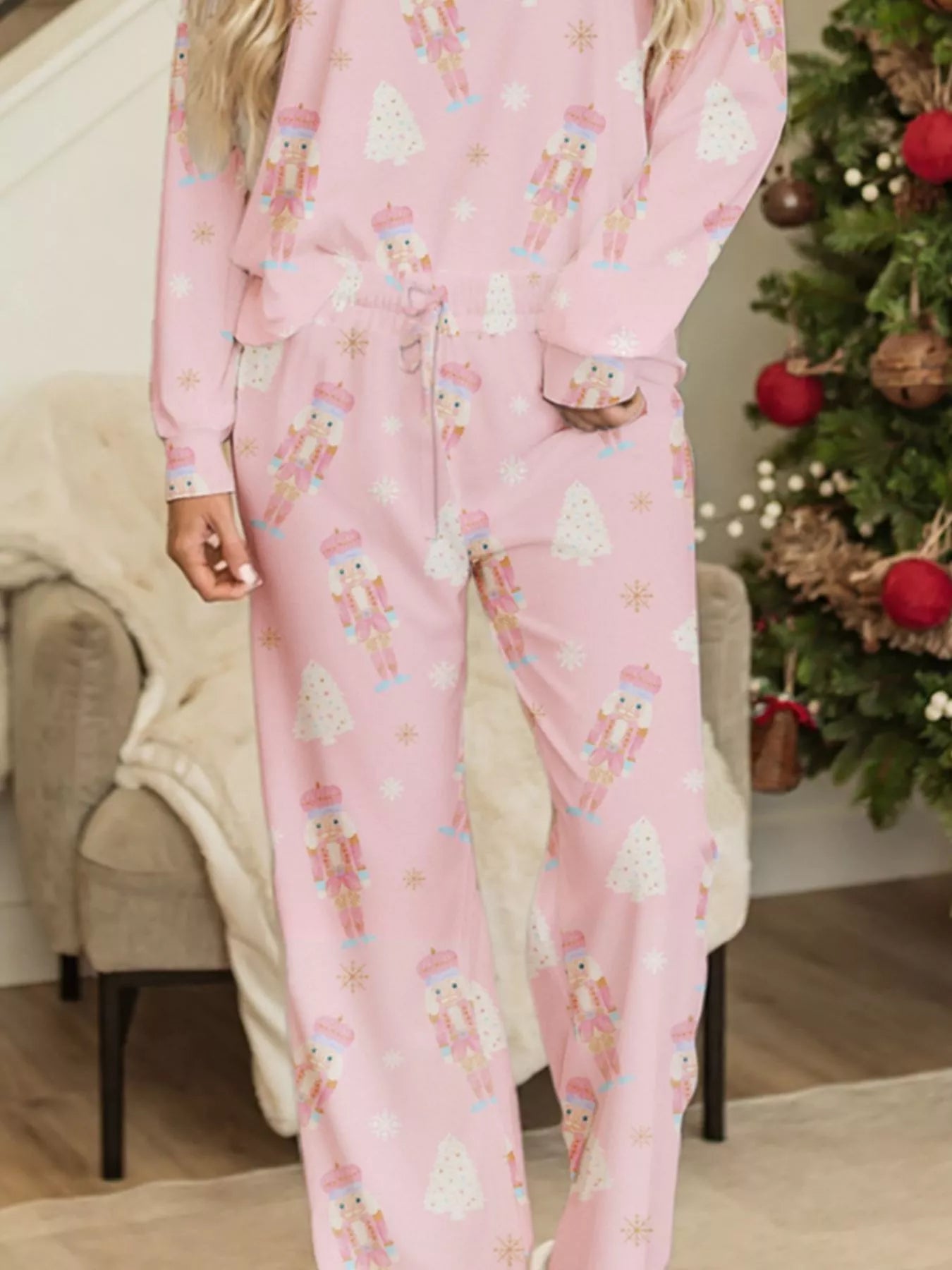 Pink two-piece nutcracker-print lounge set with long sleeves and drawstring pants worn indoors beside a Christmas tree.
