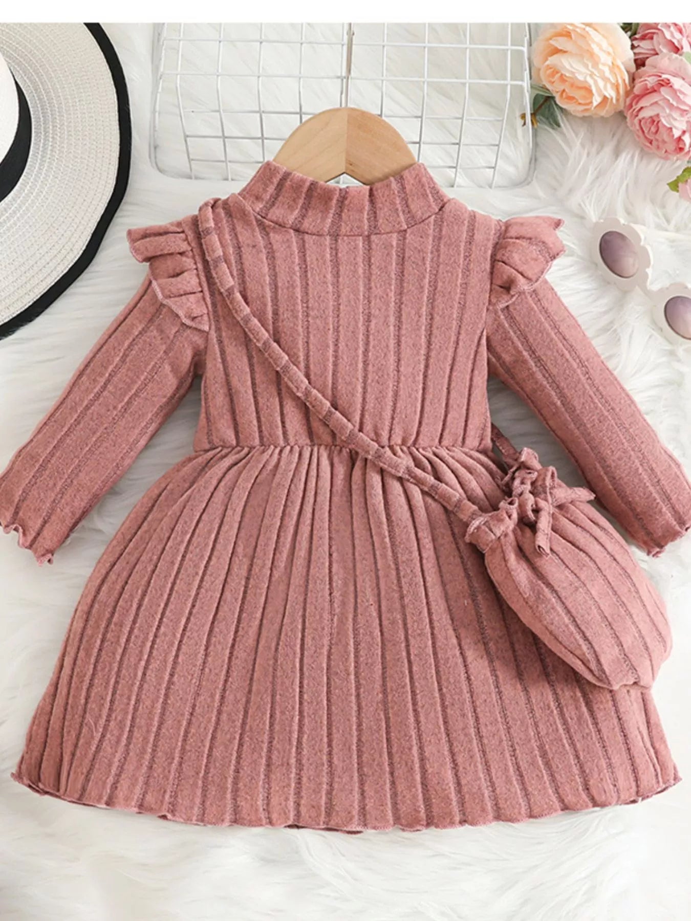 Soft pink ribbed sweater dress for girls featuring ruffled sleeves and matching purse, laid flat beside floral decor and accessories.