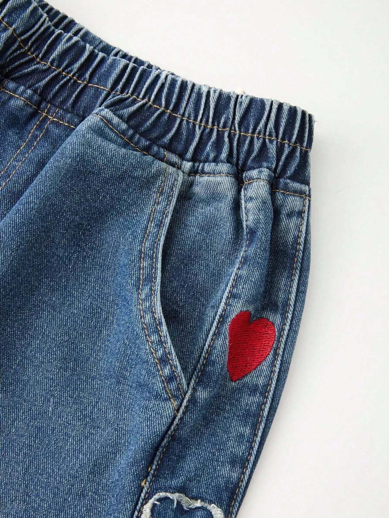 Relaxed denim pants with embroidered red and white heart details along the side panels, elastic waistband, and cuffed hems.