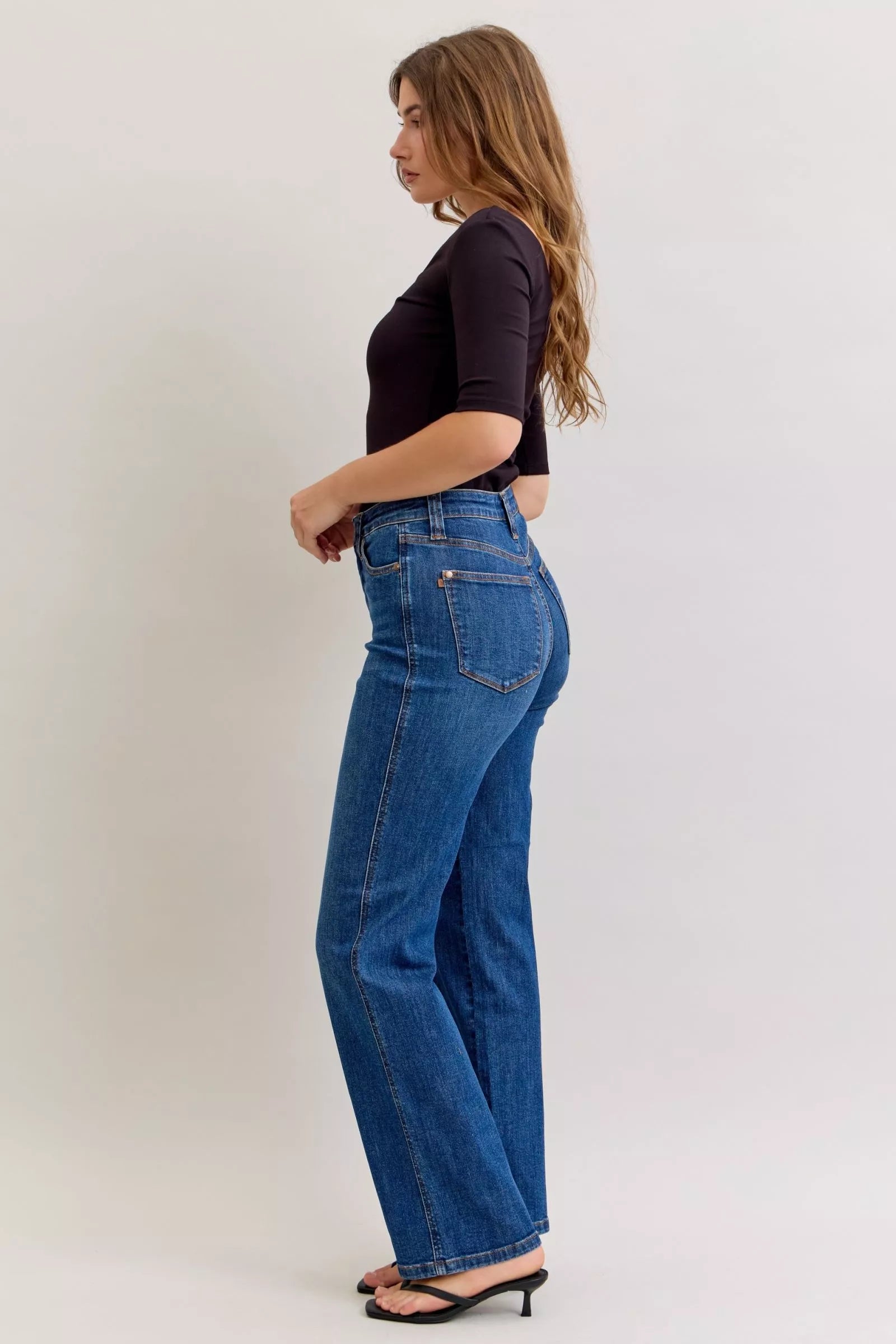 Model wearing dark wash high-rise straight jeans with tummy control fit, styled with a black top and sandals.