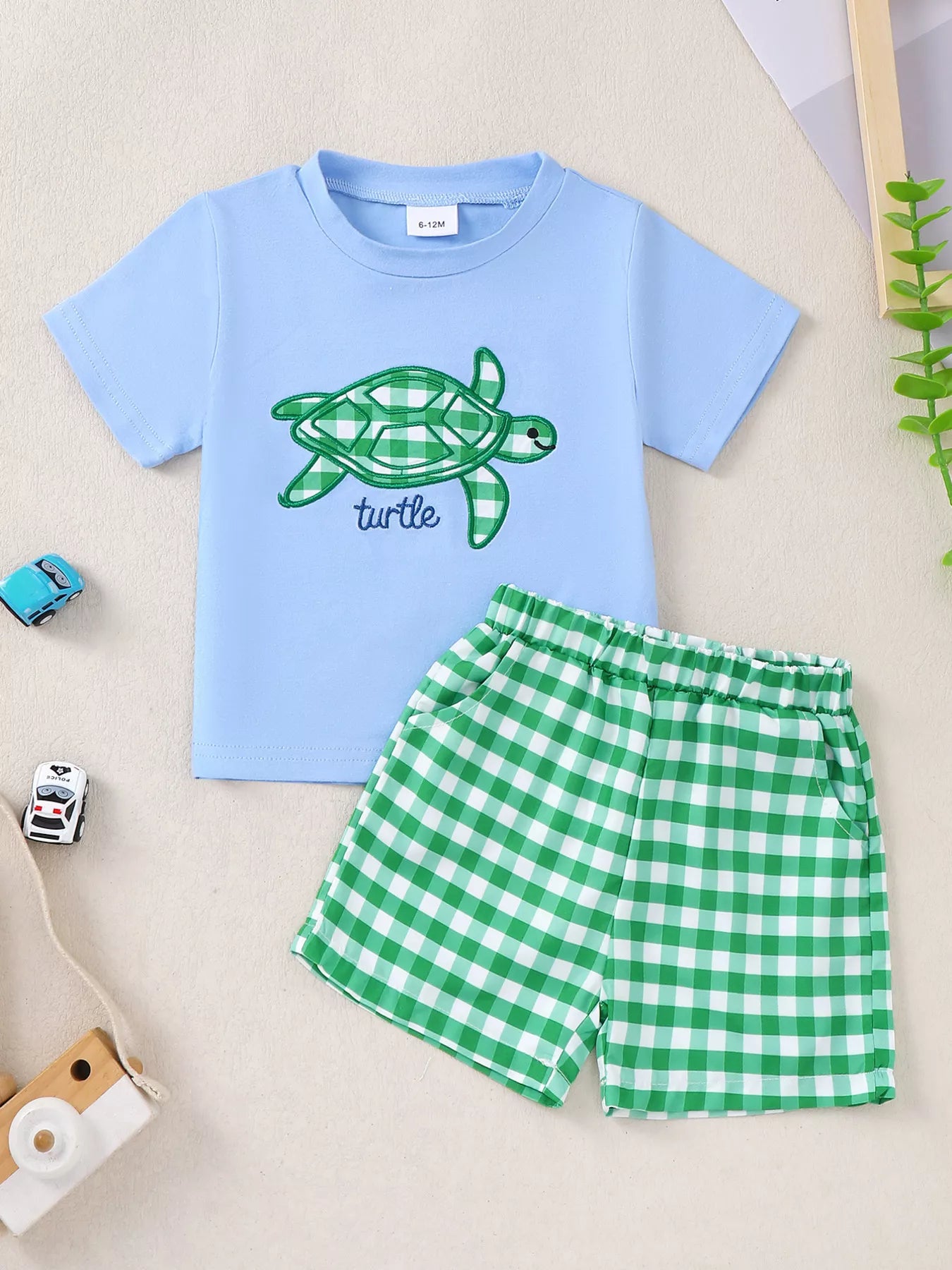 Two-piece baby outfit with blue turtle appliqué T-shirt and green gingham shorts made from a soft cotton-blend fabric.