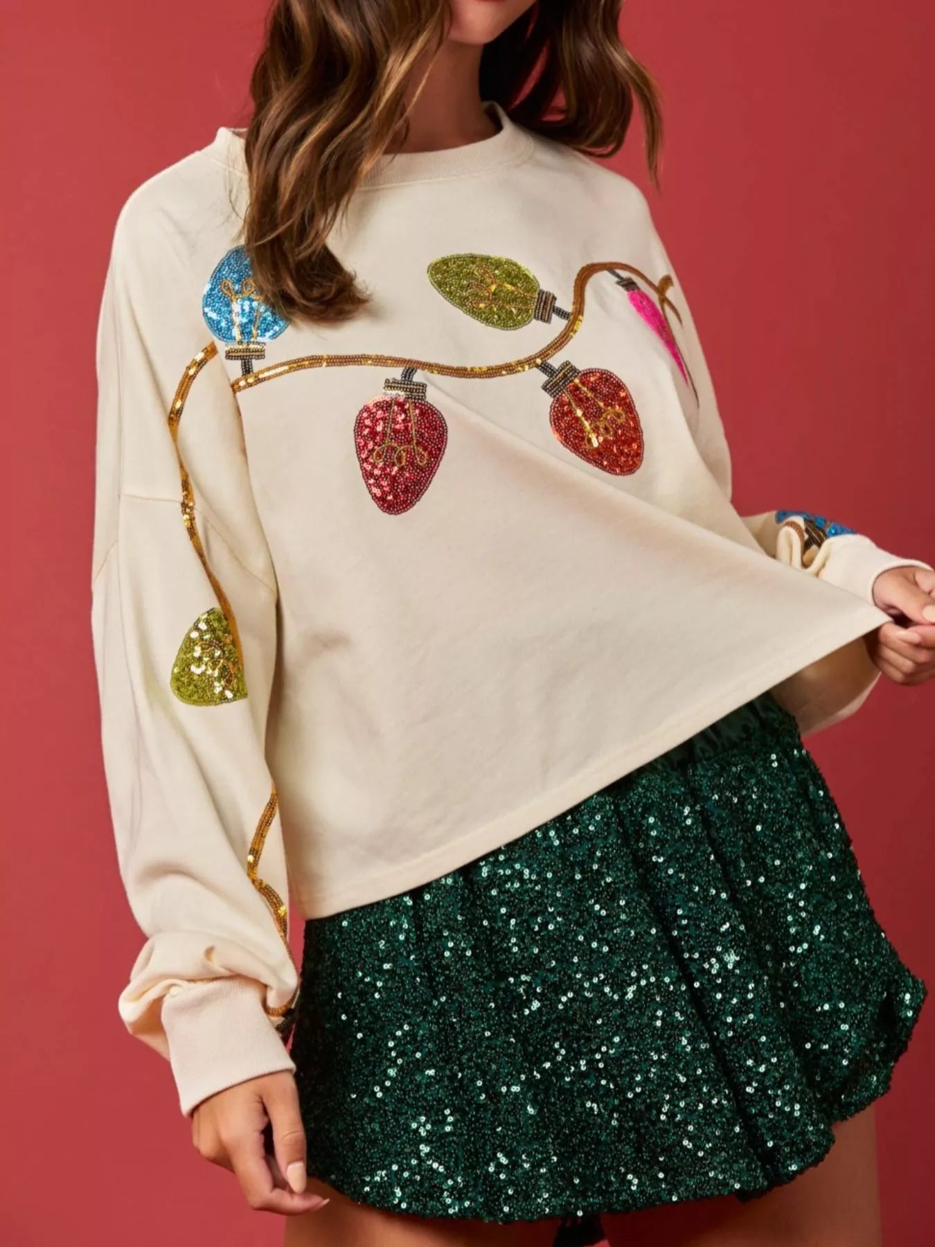 Woman wearing a cream sweatshirt with rainbow sequin Christmas lights and a sparkly green skirt — cheerful holiday outfit.