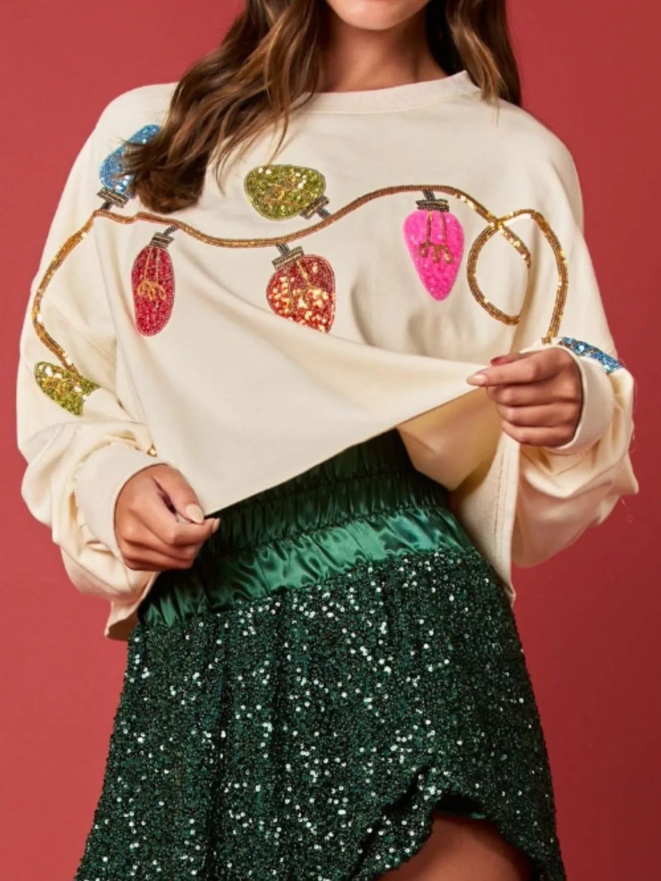 Woman wearing a cream sweatshirt with rainbow sequin Christmas lights and a sparkly green skirt — cheerful holiday outfit.