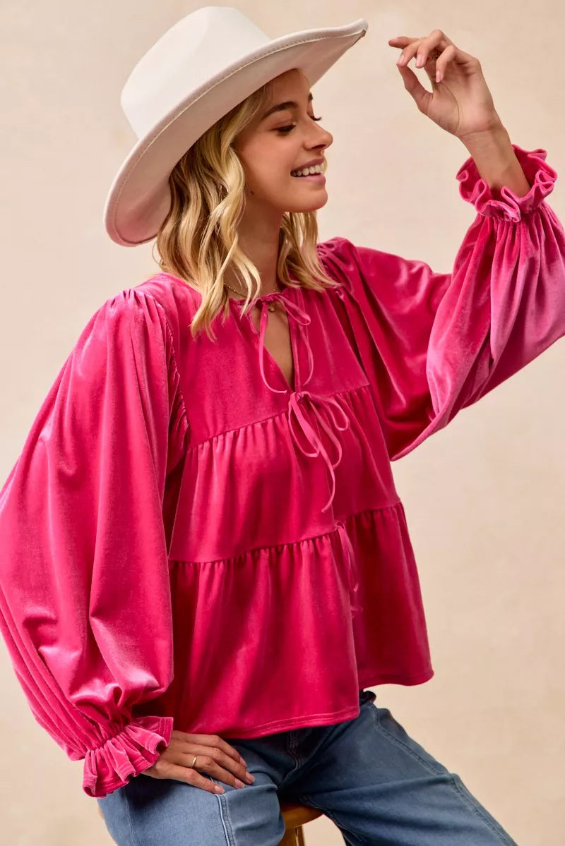 Woman wearing a fuchsia velvet long-sleeve tiered blouse with bow-tie neckline and ruffled cuffs styled with jeans and a white hat.