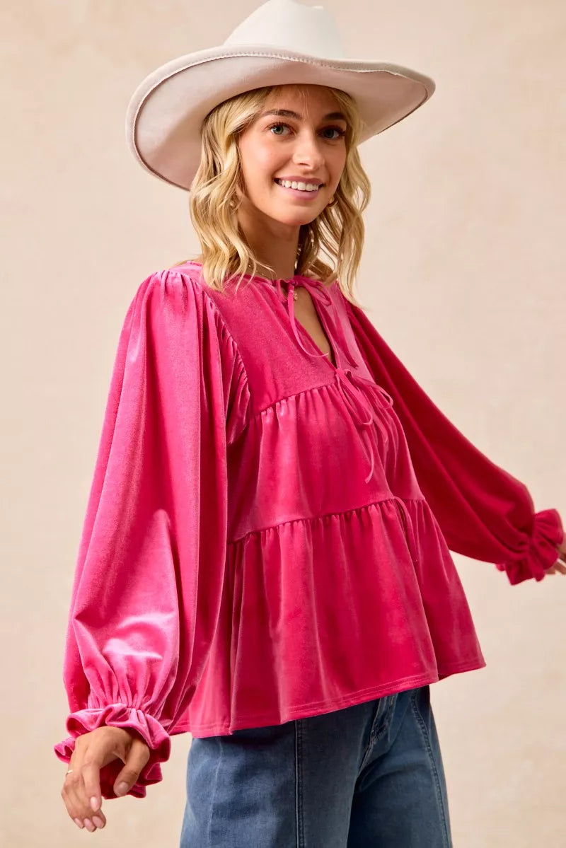 Woman wearing a fuchsia velvet long-sleeve tiered blouse with bow-tie neckline and ruffled cuffs styled with jeans and a white hat.