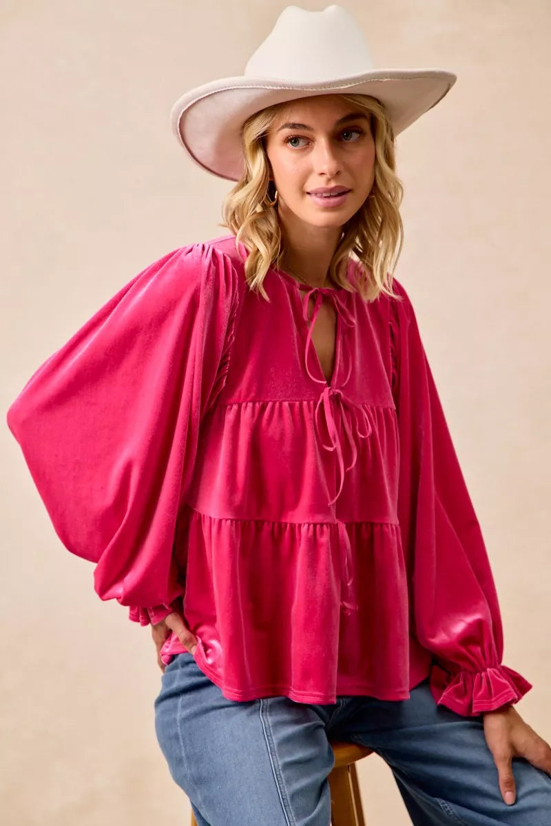 Woman wearing a fuchsia velvet long-sleeve tiered blouse with bow-tie neckline and ruffled cuffs styled with jeans and a white hat.