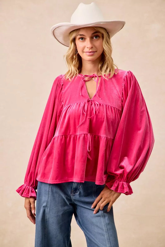 Woman wearing a fuchsia velvet long-sleeve tiered blouse with bow-tie neckline and ruffled cuffs styled with jeans and a white hat.