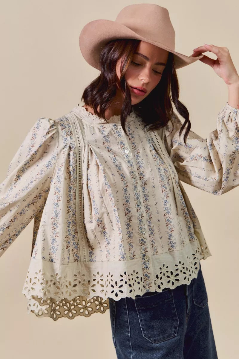 Woman wearing a cream button-down blouse with delicate floral print, lace trim, and eyelet cutout hem, styled with dark denim jeans and a tan hat.