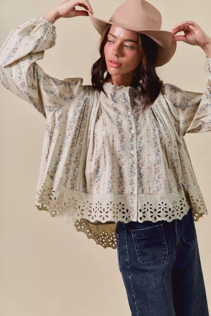 Woman wearing a cream button-down blouse with delicate floral print, lace trim, and eyelet cutout hem, styled with dark denim jeans and a tan hat.