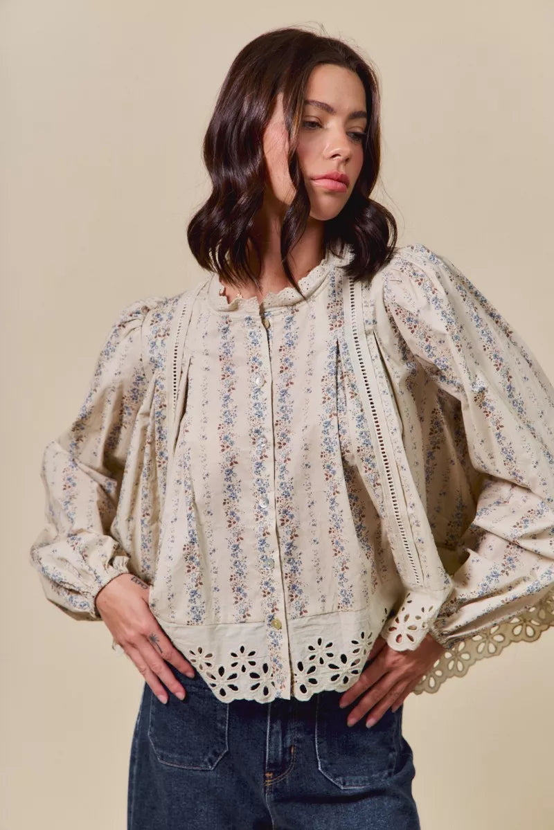 Woman wearing a cream button-down blouse with delicate floral print, lace trim, and eyelet cutout hem, styled with dark denim jeans and a tan hat.