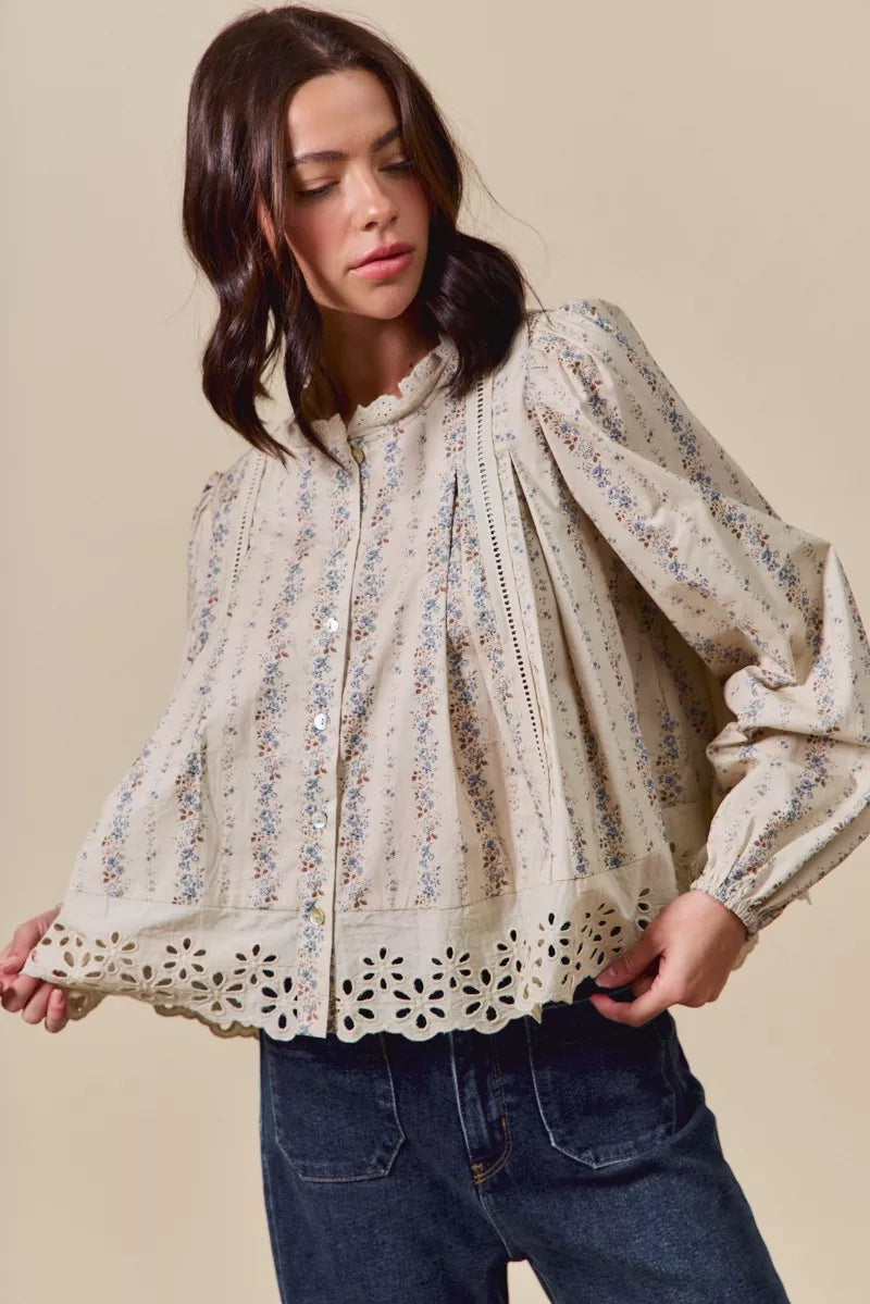 Woman wearing a cream button-down blouse with delicate floral print, lace trim, and eyelet cutout hem, styled with dark denim jeans and a tan hat.
