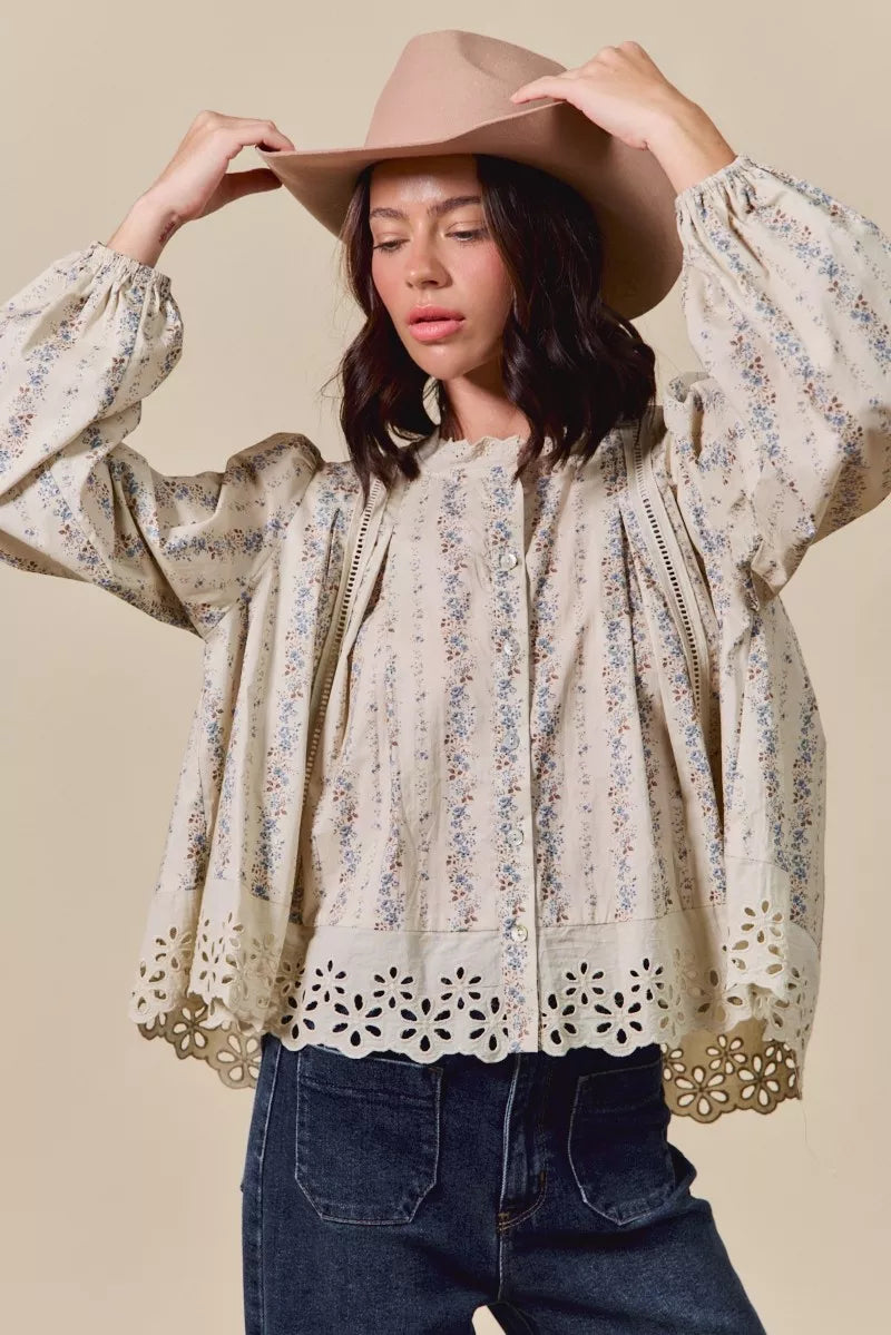 Woman wearing a cream button-down blouse with delicate floral print, lace trim, and eyelet cutout hem, styled with dark denim jeans and a tan hat.